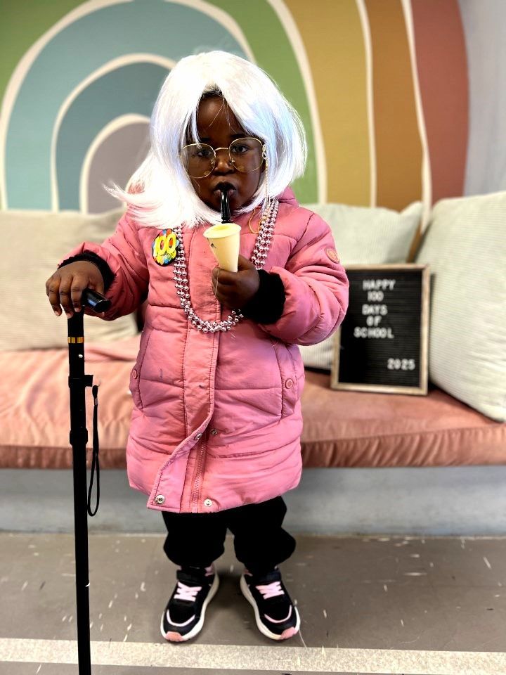 Child in pink coat and glasses, dressed as an elderly person, holding a drink and cane.