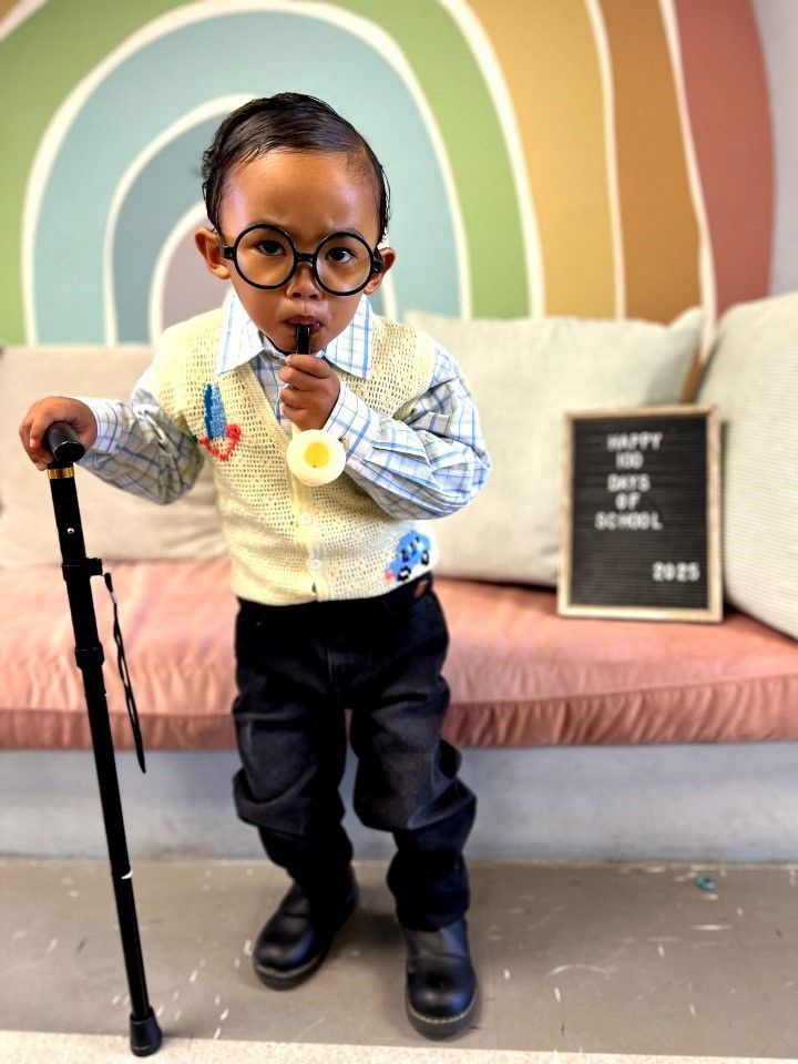 Child dressed as a 100-year-old for school, holding a cane and lollipop, wearing glasses and a vest.