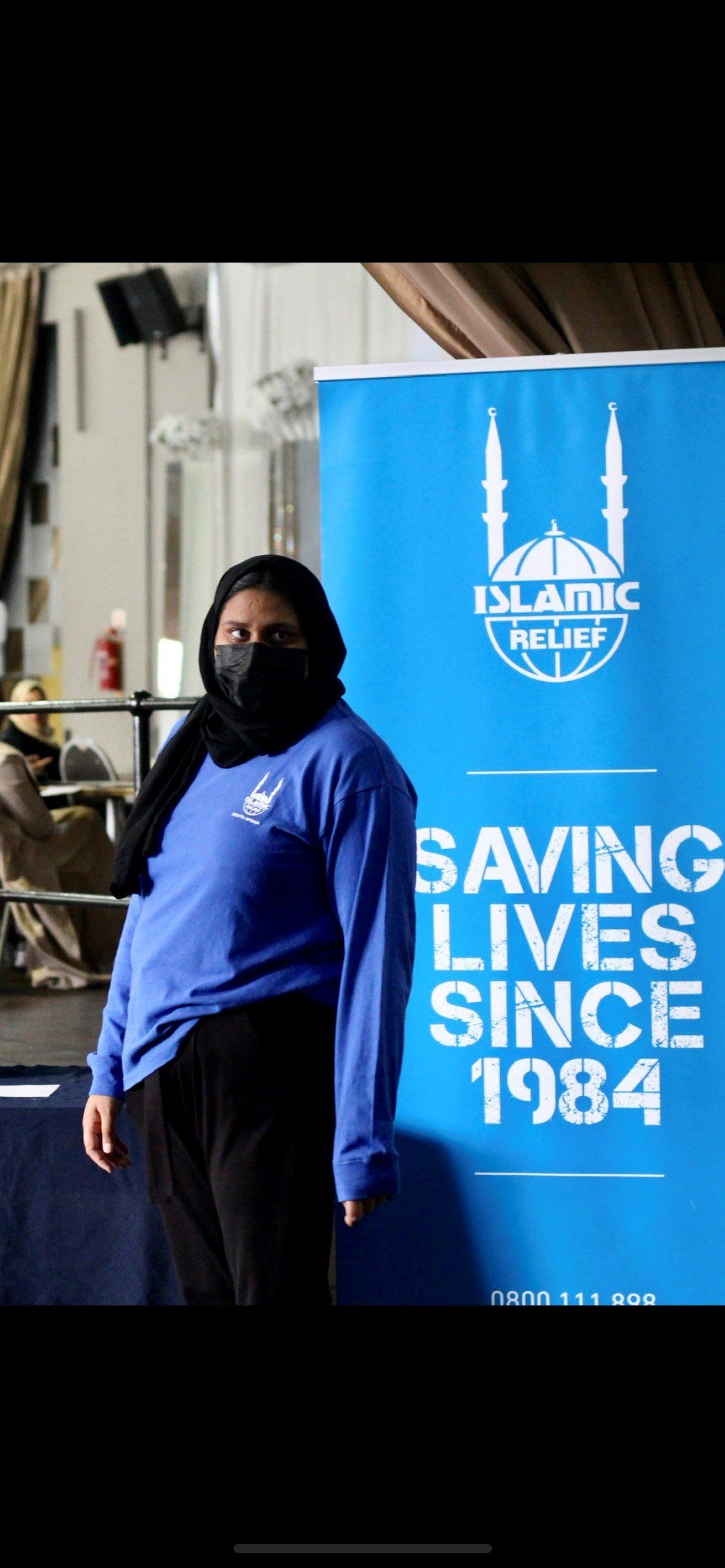 A woman wearing a mask is standing in front of a sign that says saving lives since 1984.