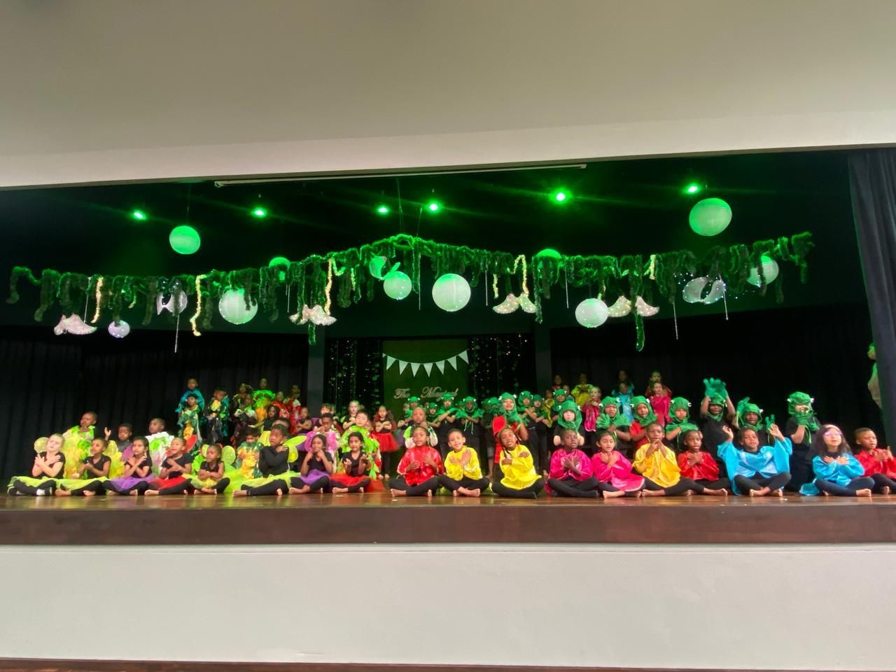 A group of children are sitting on a stage with green lights hanging from the ceiling.