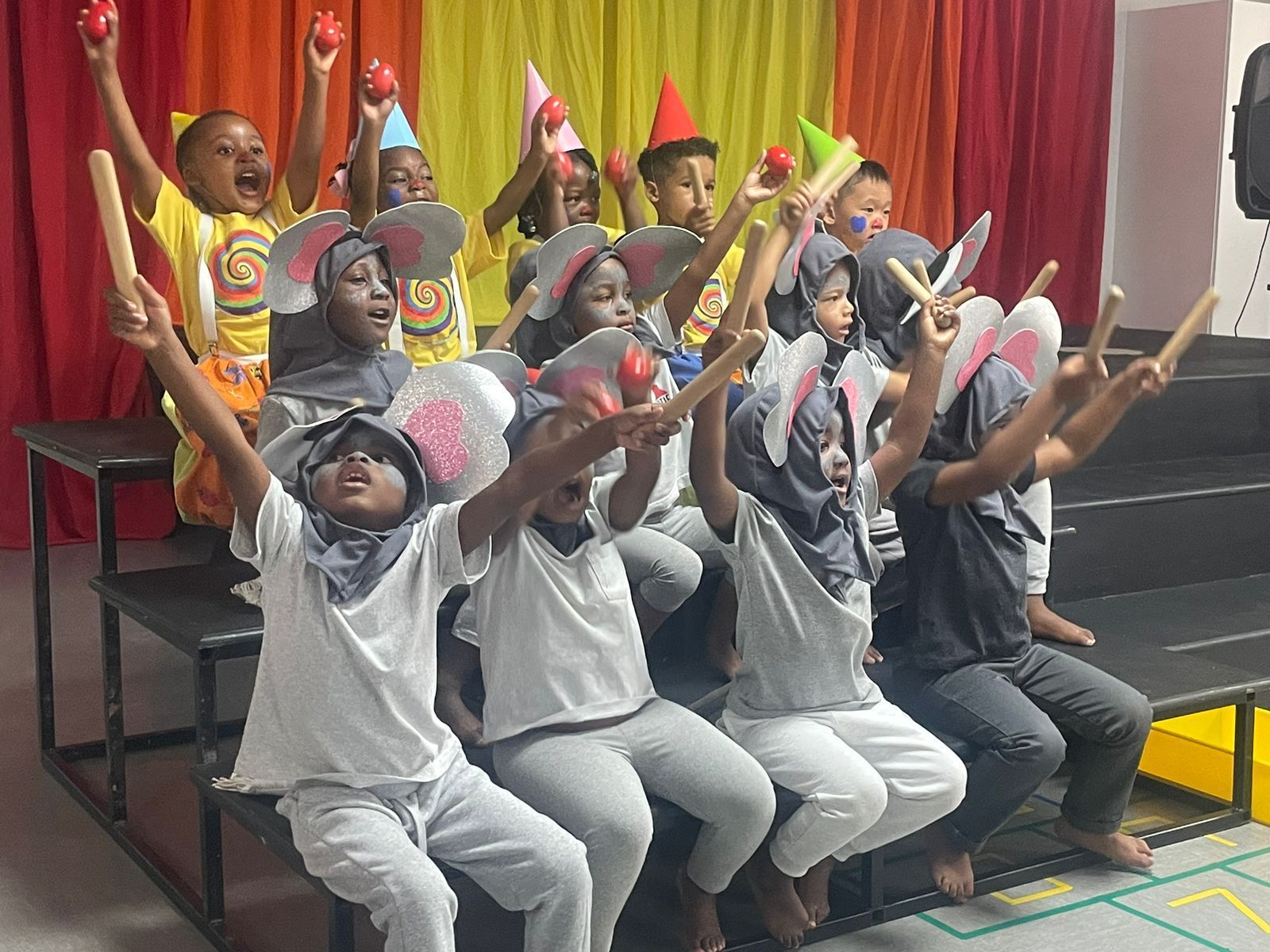 A group of children are sitting on a stage with their arms in the air.