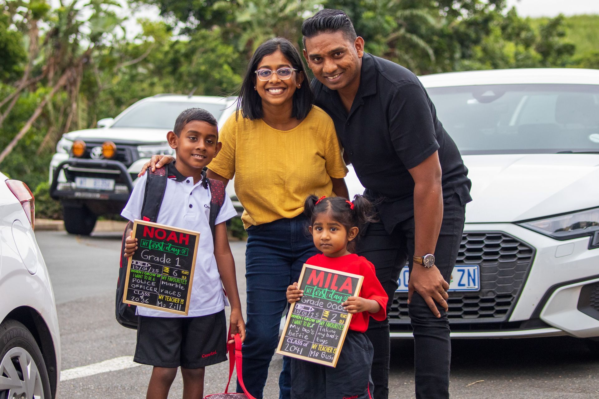 Family poses with children holding signs, near cars.