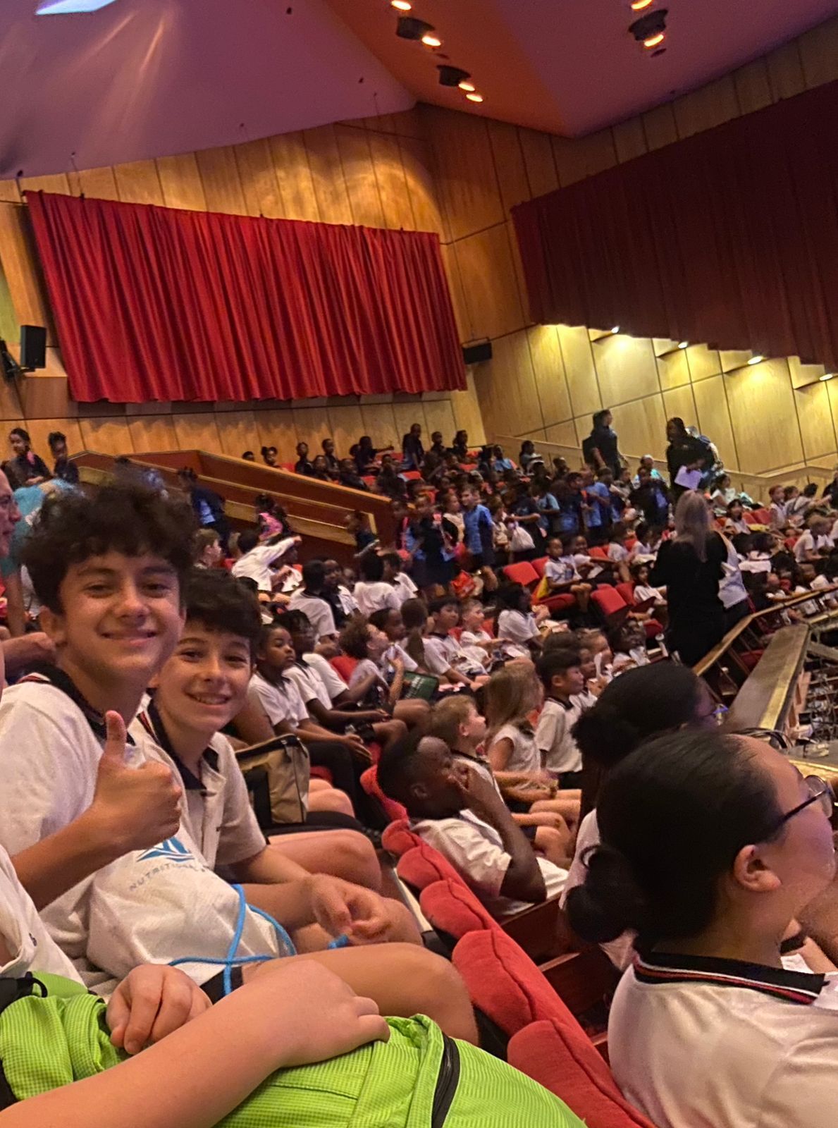 A group of children are sitting in an auditorium giving thumbs up.