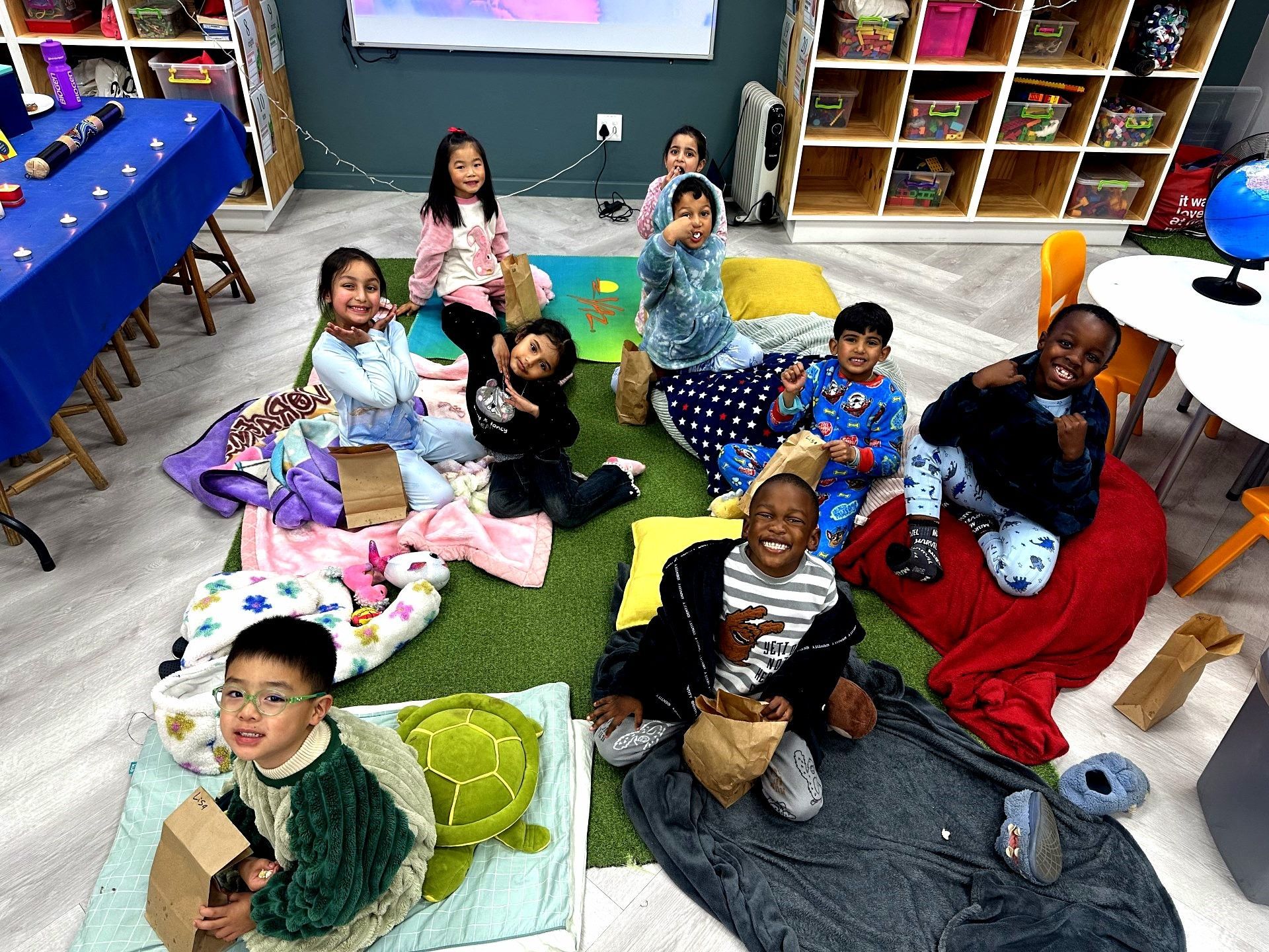 Children in pajamas sit on blankets, eating snacks, in a brightly lit classroom.