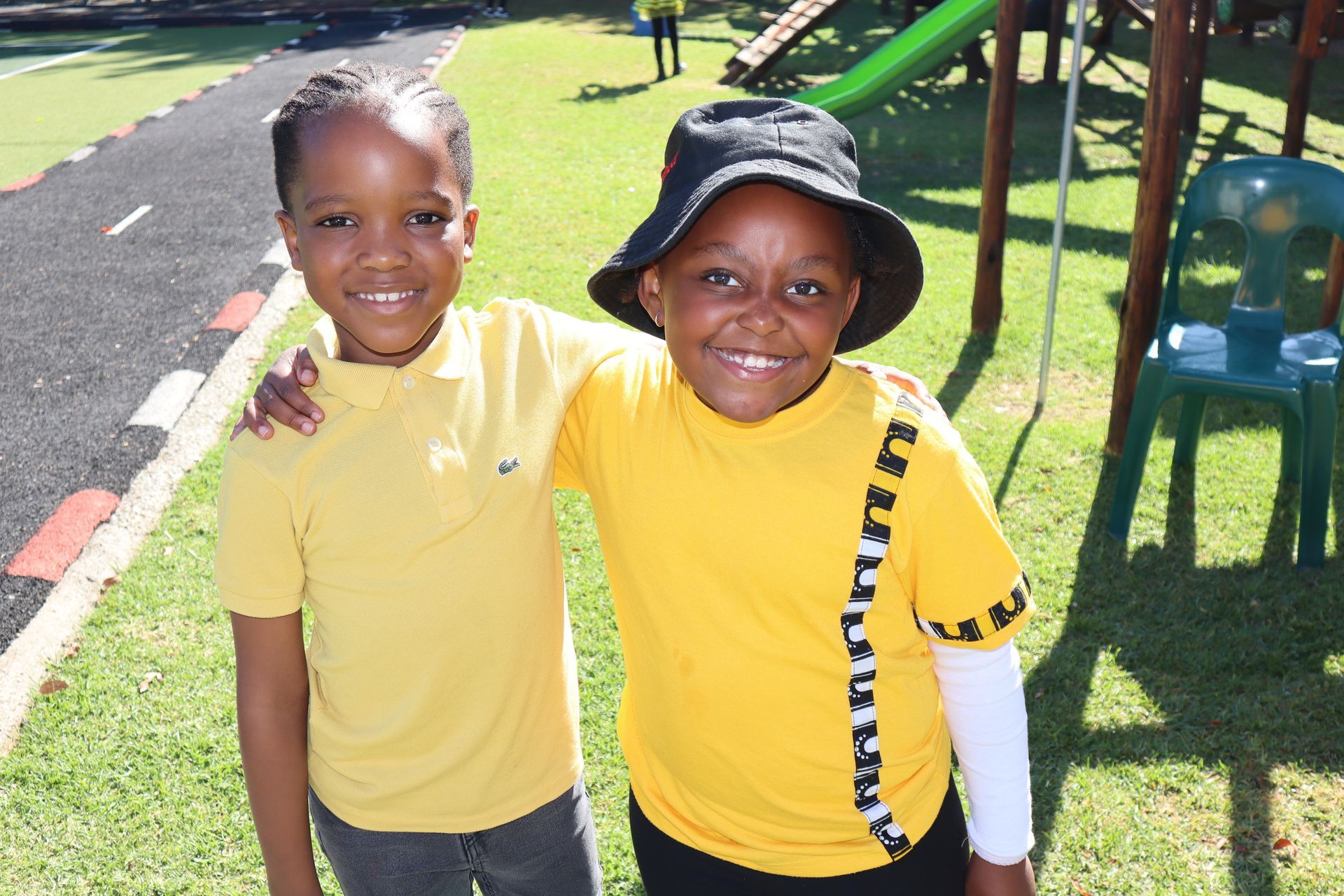 Two children in yellow shirts smile, one with an arm around the other, near a playground.