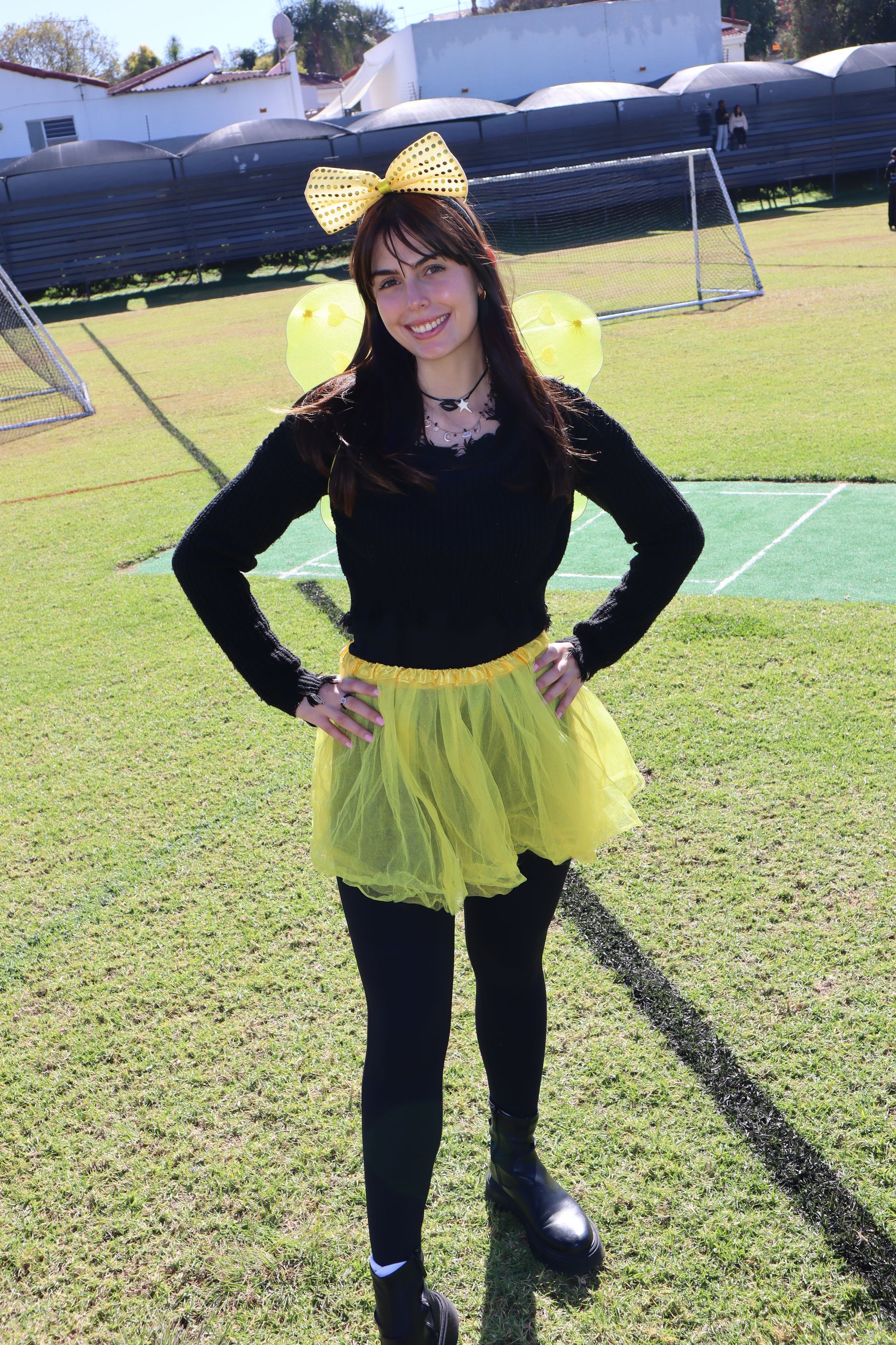 Woman in a bee costume stands on a green field. She wears a black top, yellow tutu, and bee antenna.