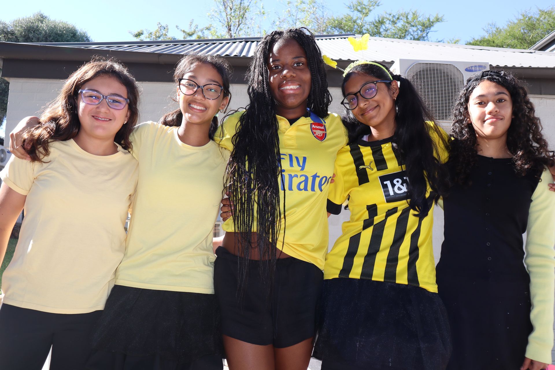 Five smiling people in yellow shirts, black skirts, and glasses pose outdoors, arms around each other.