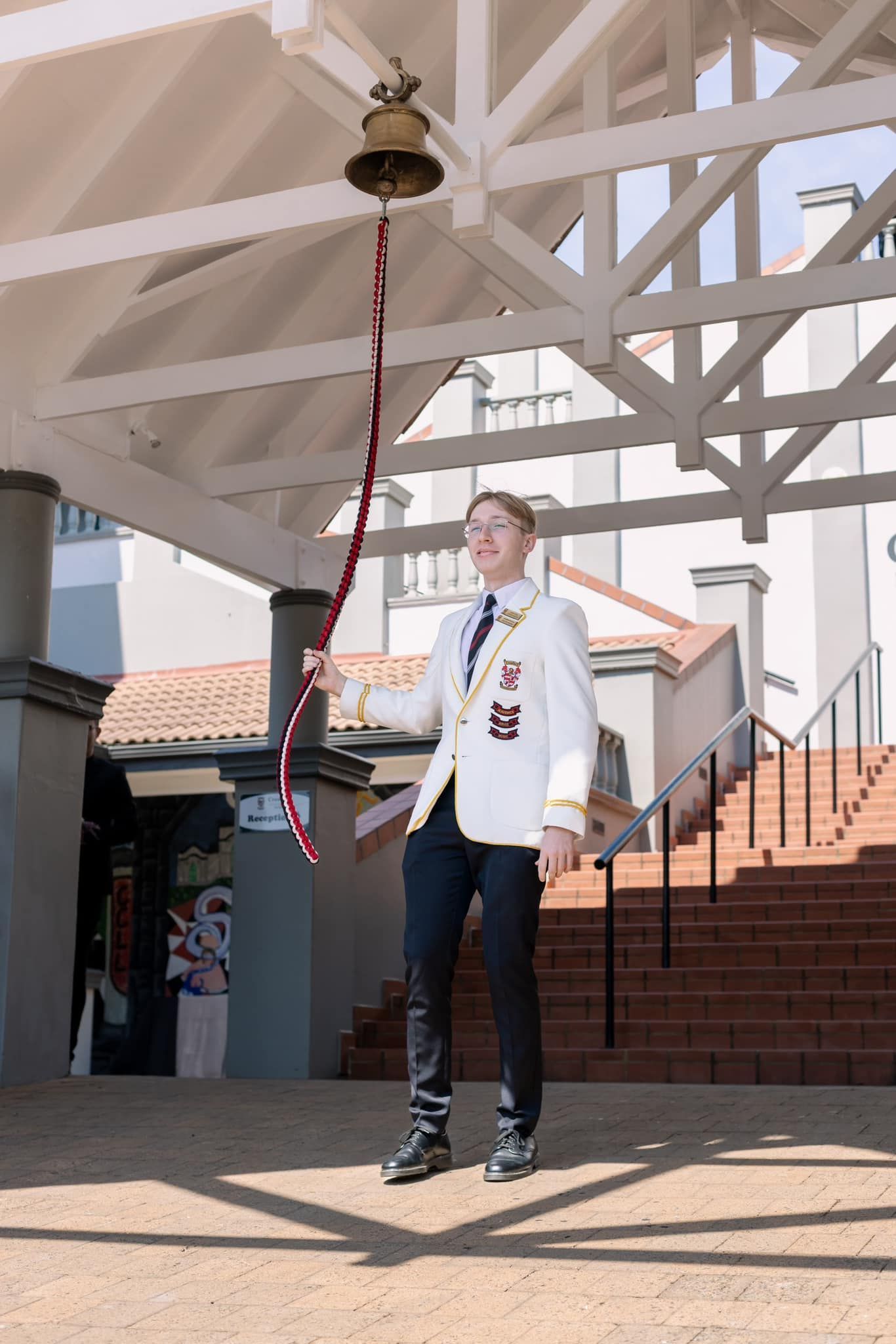 A man in a white jacket is standing under a roof holding a bell.