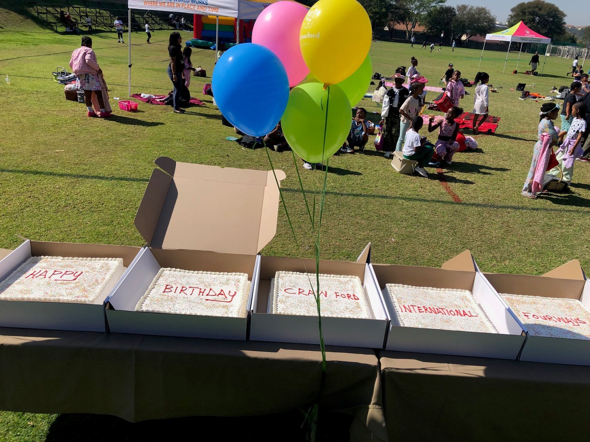 A bunch of balloons are hanging from a string next to some boxes of birthday cakes