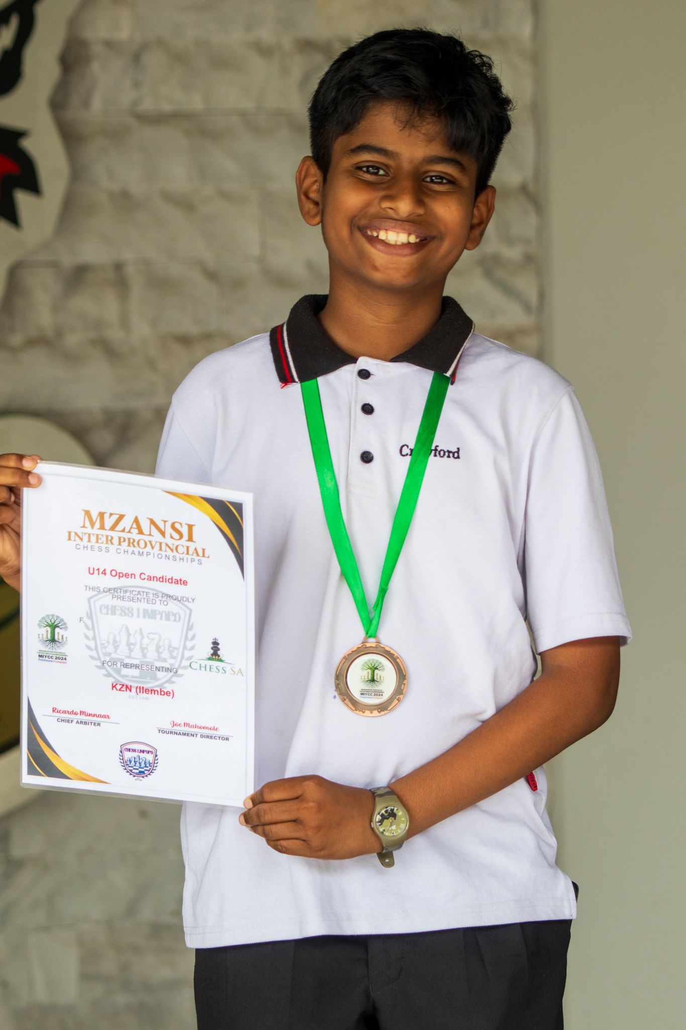 Boy in white shirt with medal holds a certificate, smiling outside.