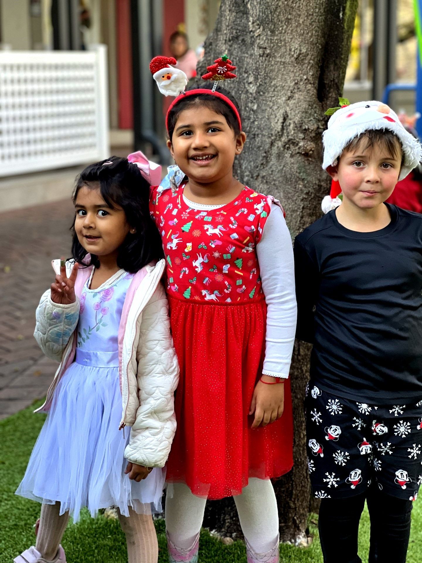 Three children dressed for Christmas, standing outdoors by a tree; one wears a Santa hat and pajamas.