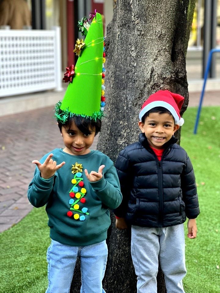 Two children, one wearing a Christmas tree hat and the other a Santa hat, standing next to a tree.