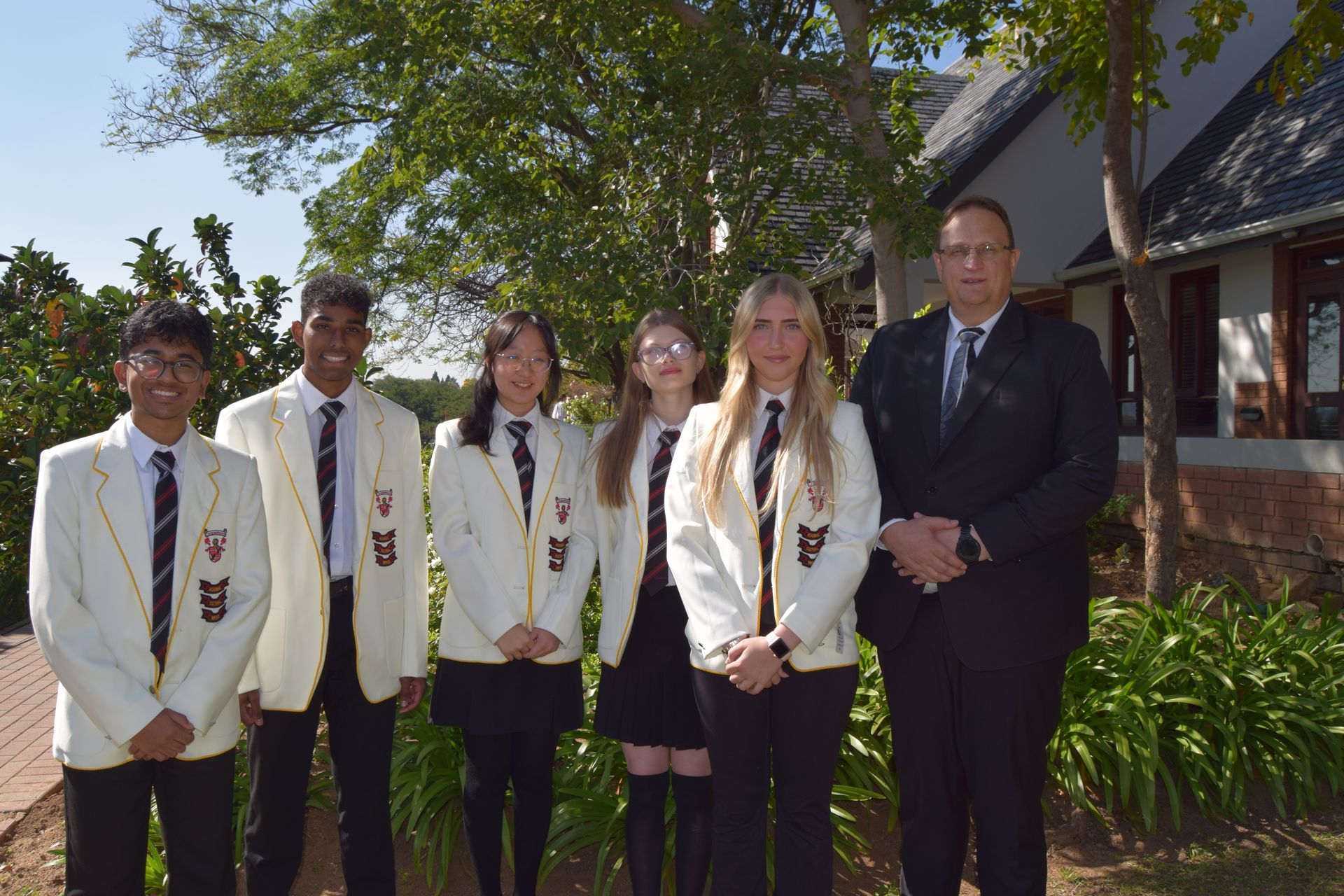 Six people, including a man in a suit, pose outdoors in front of a building and trees. Students in blazers.