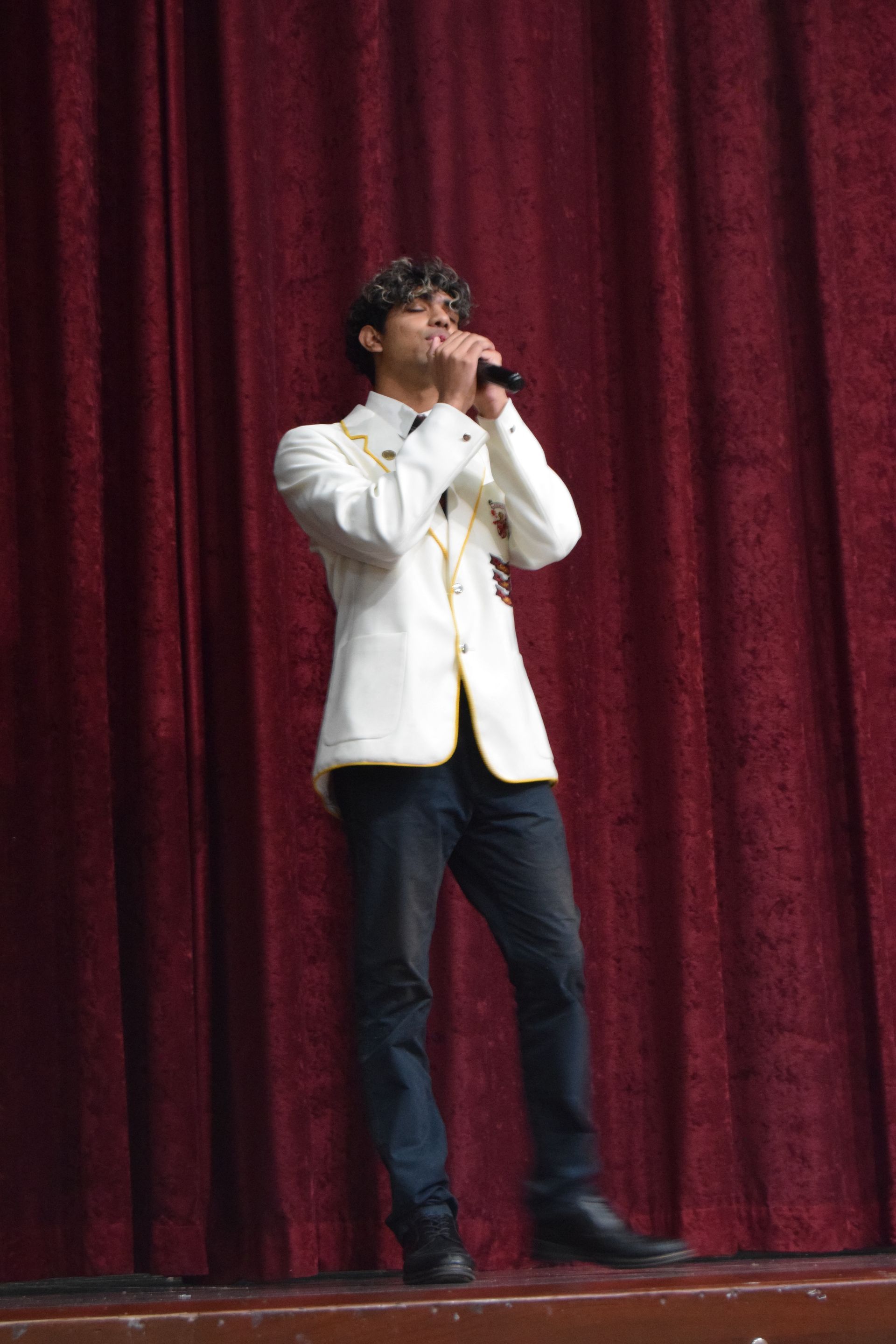 Man in a white blazer and jeans sings into a microphone on a stage with red curtains.