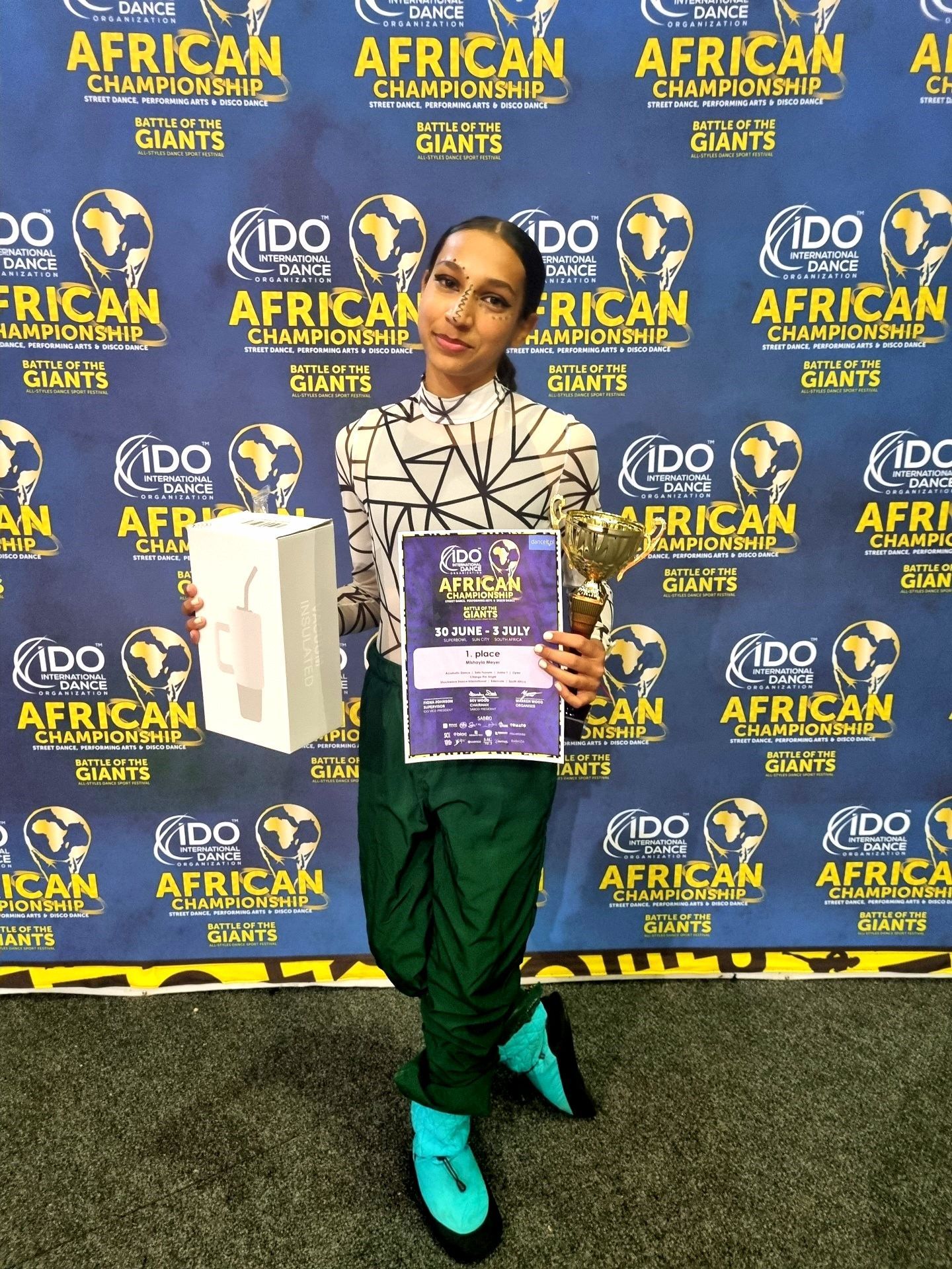 Young person holding awards, certificate, and box, in front of an event backdrop with logos.