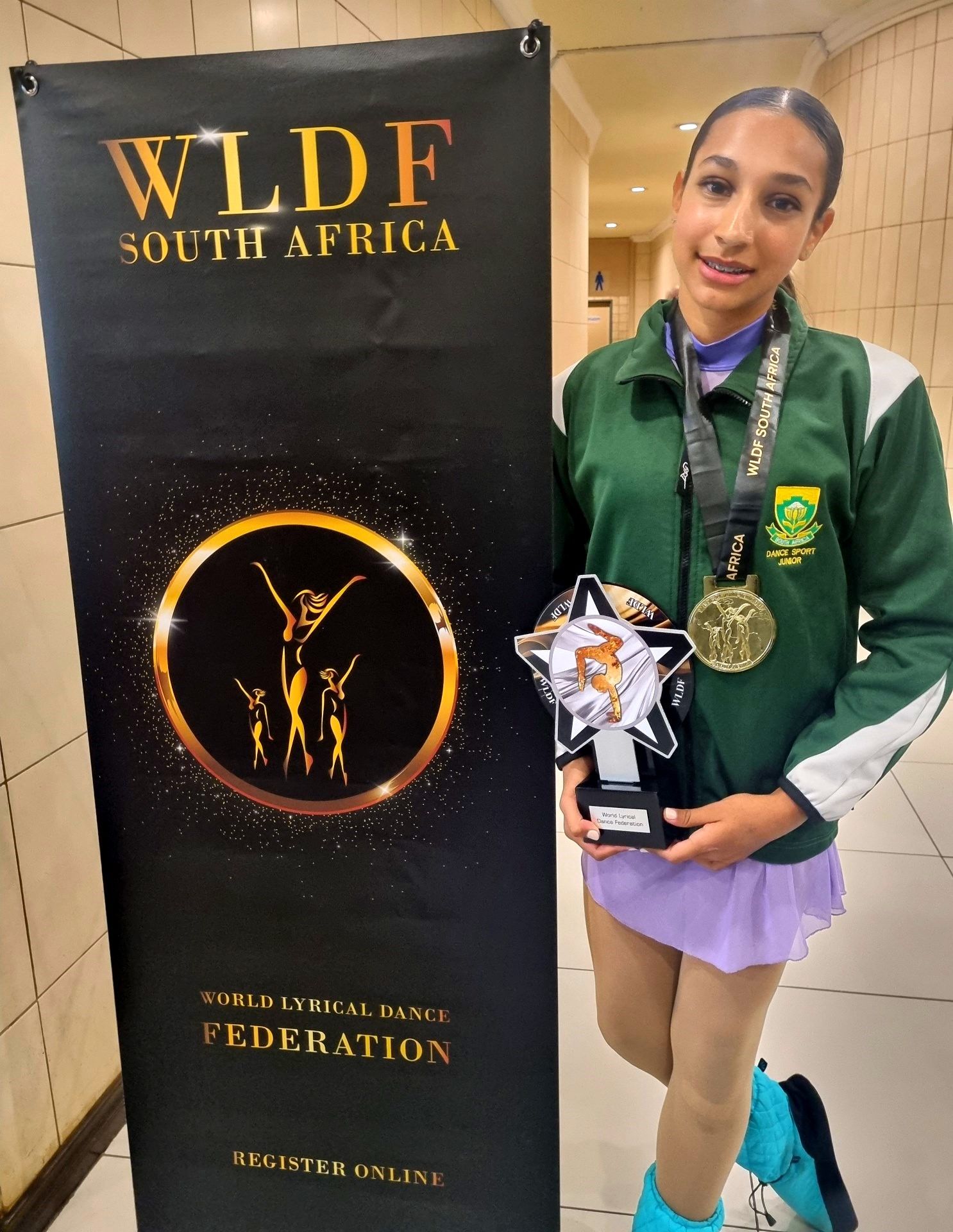 Young person in green jacket, ice skating outfit, holding trophy and medal beside a WLDFA banner.