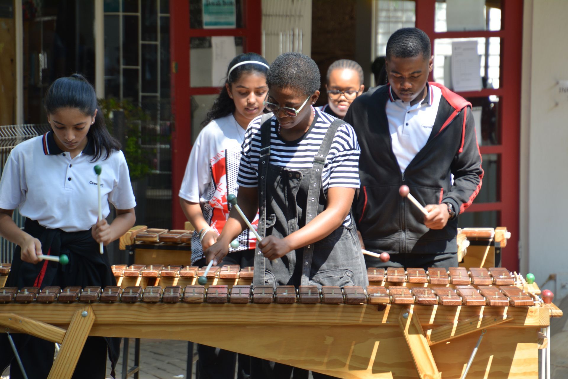 A group of children are playing xylophones in front of a red door.