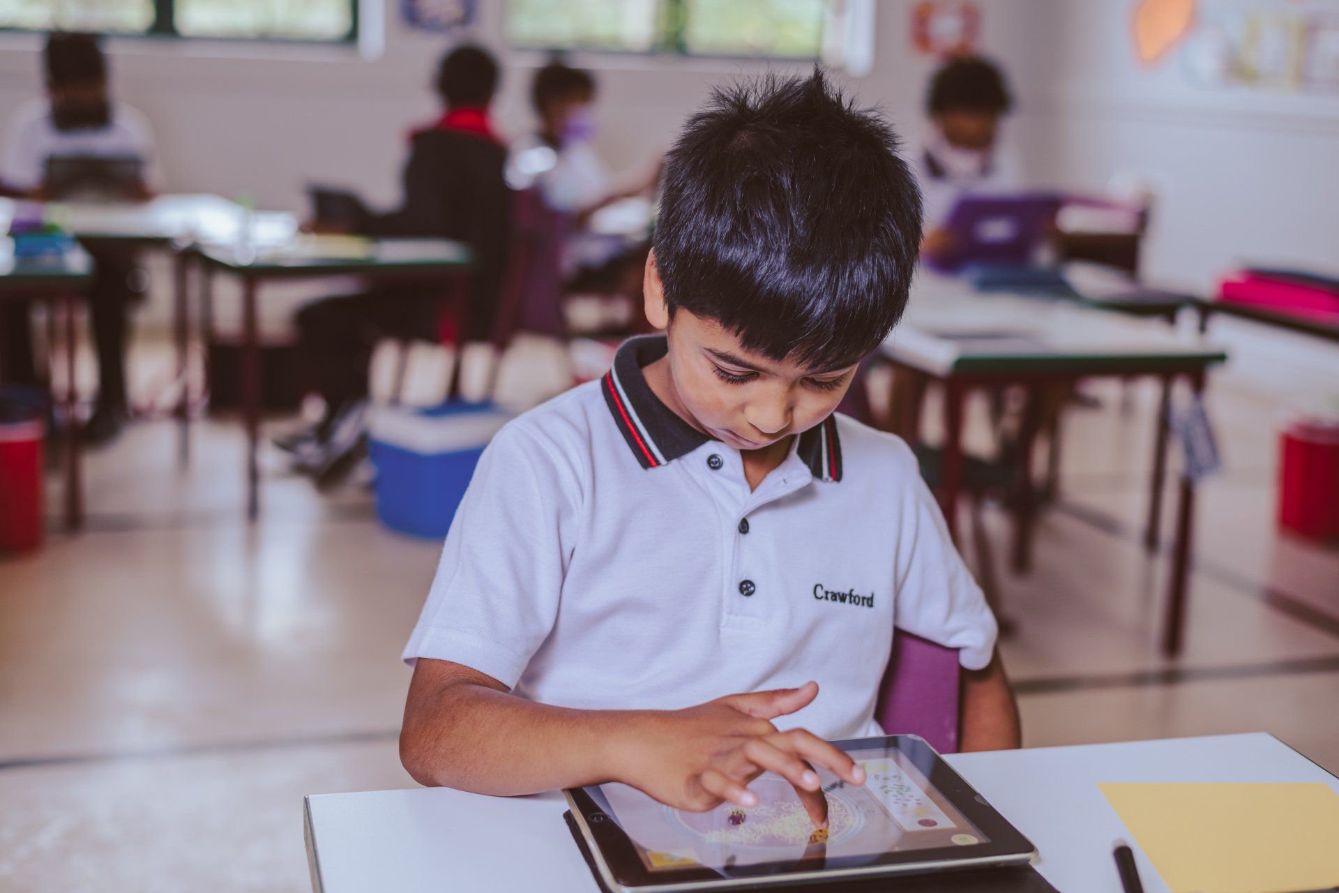 a young boy is sitting at a desk in a classroom using a tablet computer .