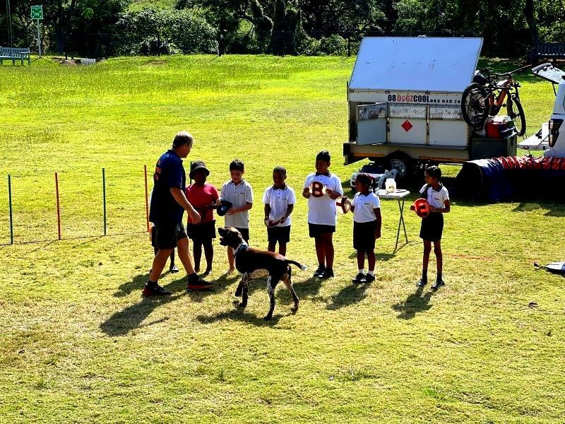 Dog training lesson with children on a grassy field. A trainer, a dog, and seven kids are present.