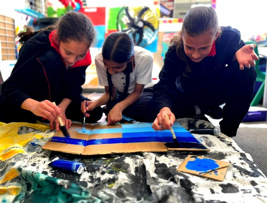 Three children painting blue stripes on cardboard, focused expressions, art project.