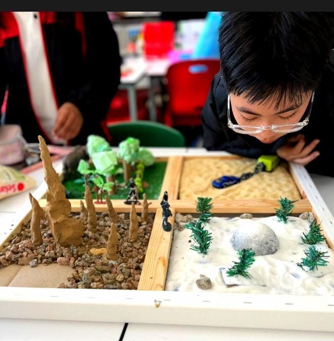 Boy with glasses examines a miniature landscape diorama with sand, trees, and rocks in a classroom.