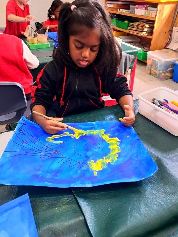 Girl painting with a brush, creating yellow shapes on a blue paper at a table. Classroom setting.