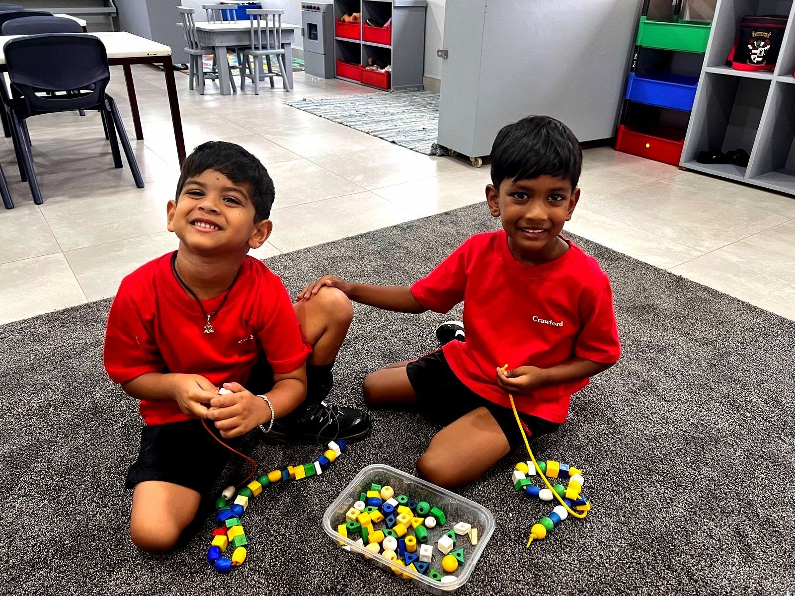 Two children stringing beads on a rug in a classroom, smiling. They wear red shirts and black shorts.