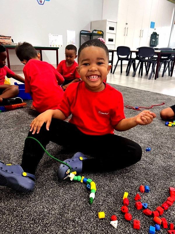 Smiling child in red shirt and black pants sits on rug, stringing beads. Others play nearby.