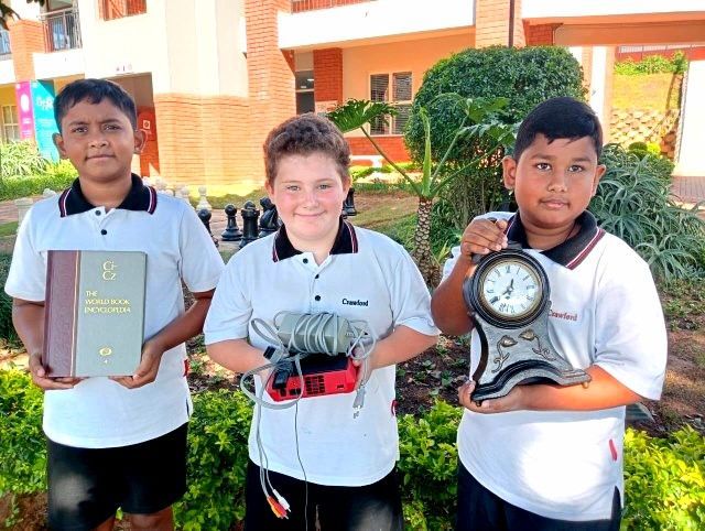 Three students in school uniforms holding items: a book, electronics, and a clock outdoors.
