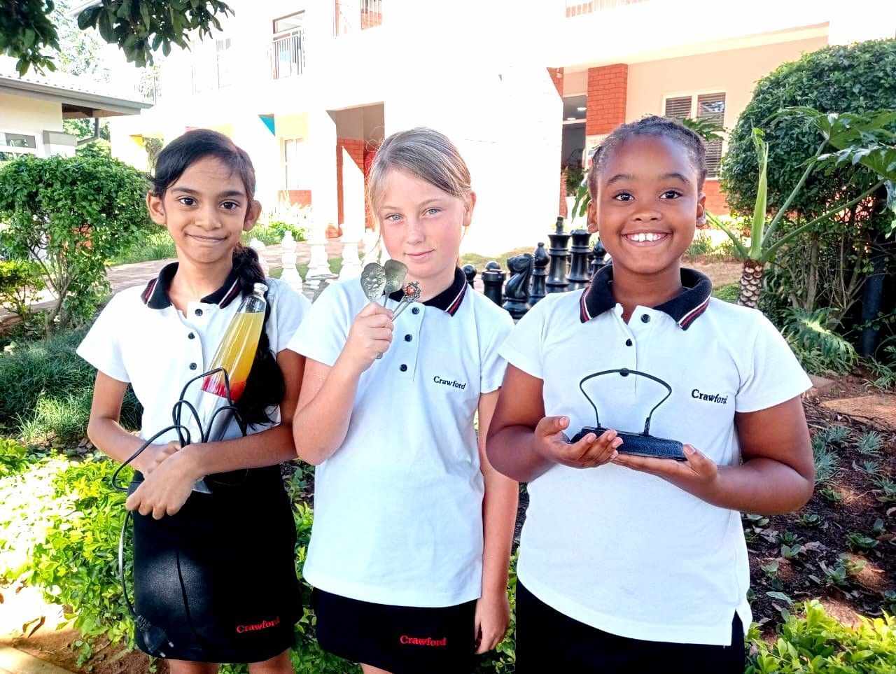 Three students holding science project components outdoors, smiling.