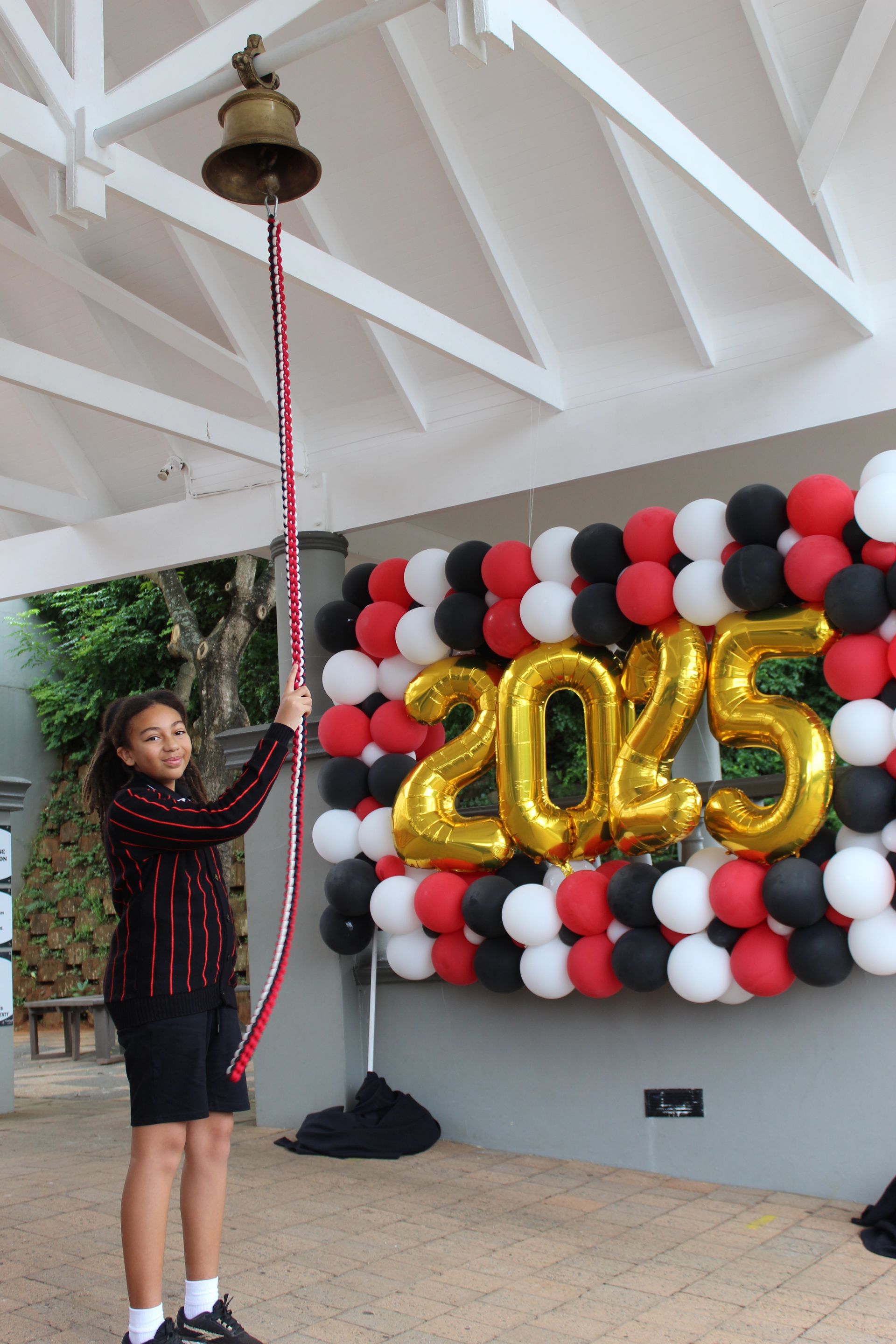 Girl ringing a bell under a white pergola. Balloons behind her read 