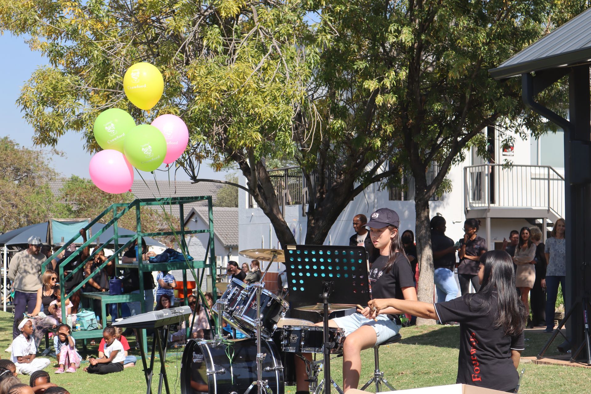 A group of people are playing instruments in a park with balloons hanging from a tree.