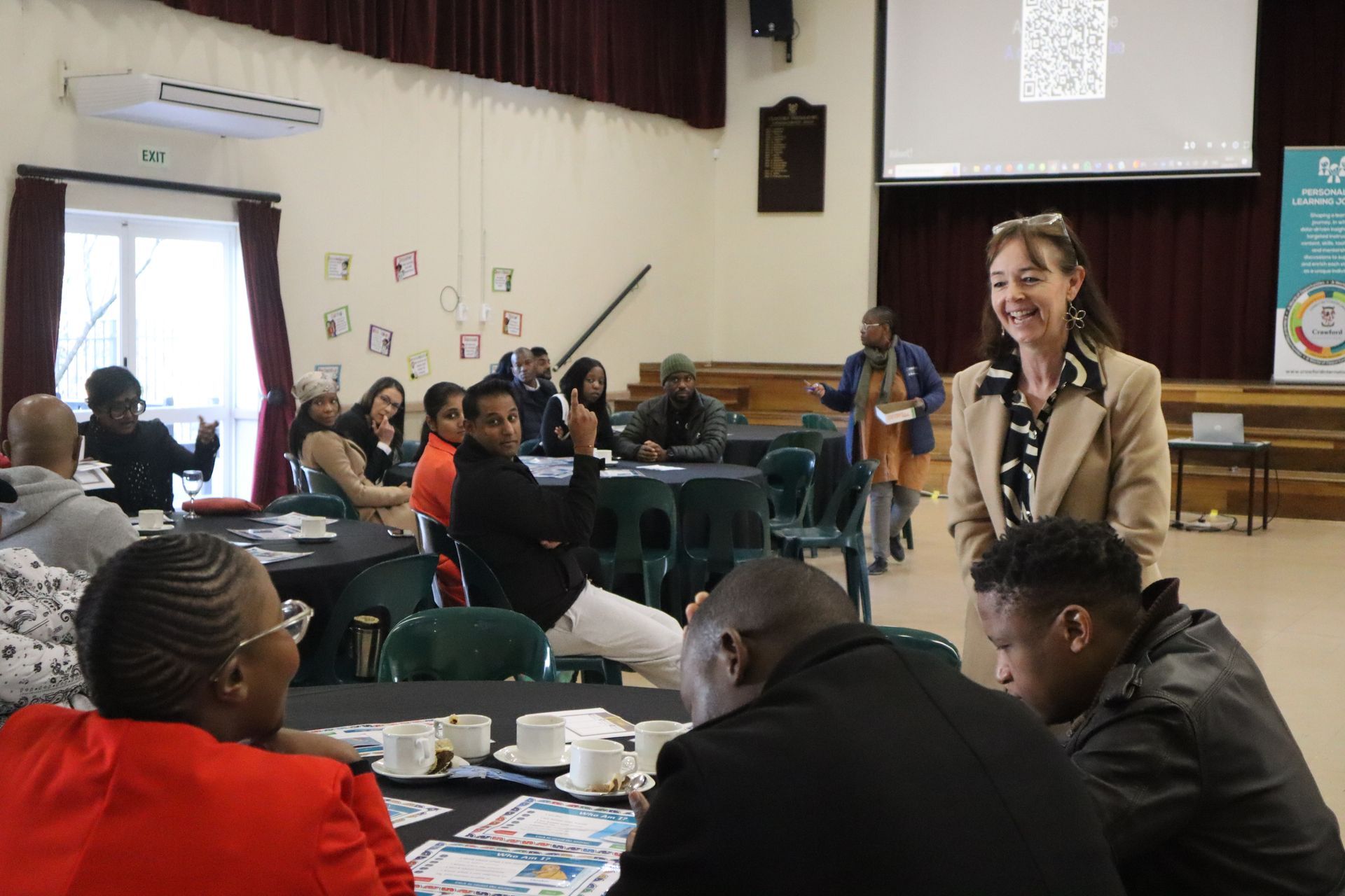 A woman is standing in front of a group of people sitting at tables in a room.