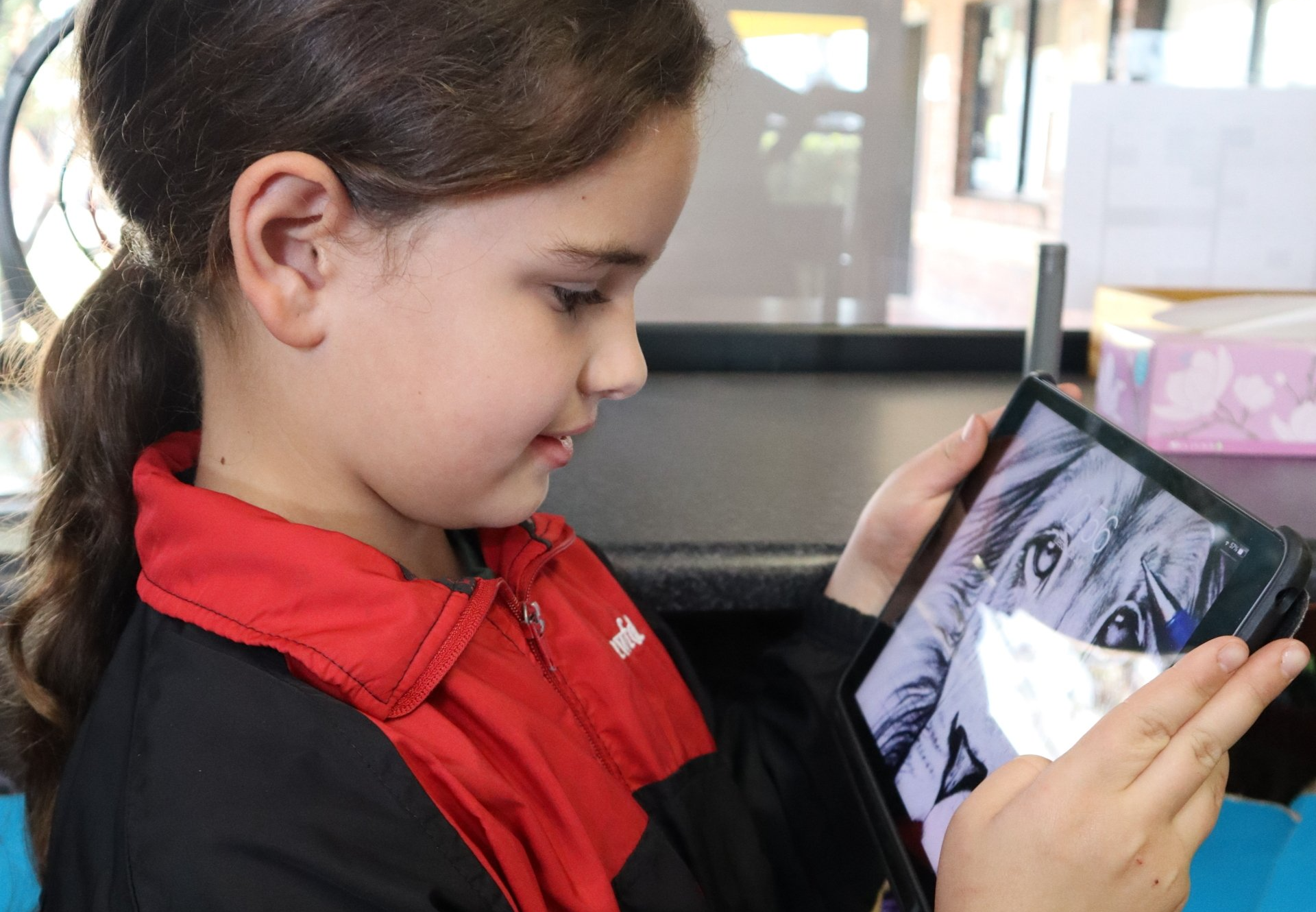A little girl in a red jacket is looking at a tablet.