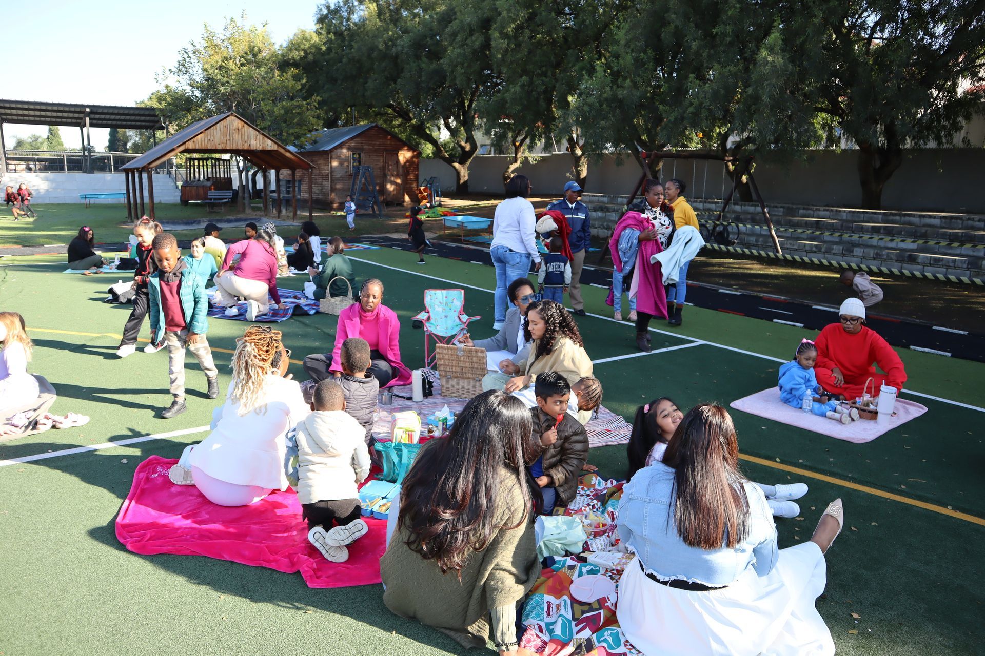 People picnic on a grassy field; blankets, food, and a small wooden structure in the background.