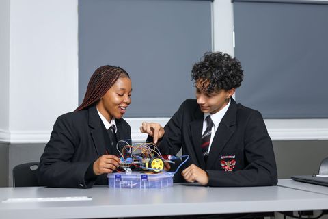 Two students in uniform work on a robotics project at a desk, smiling.