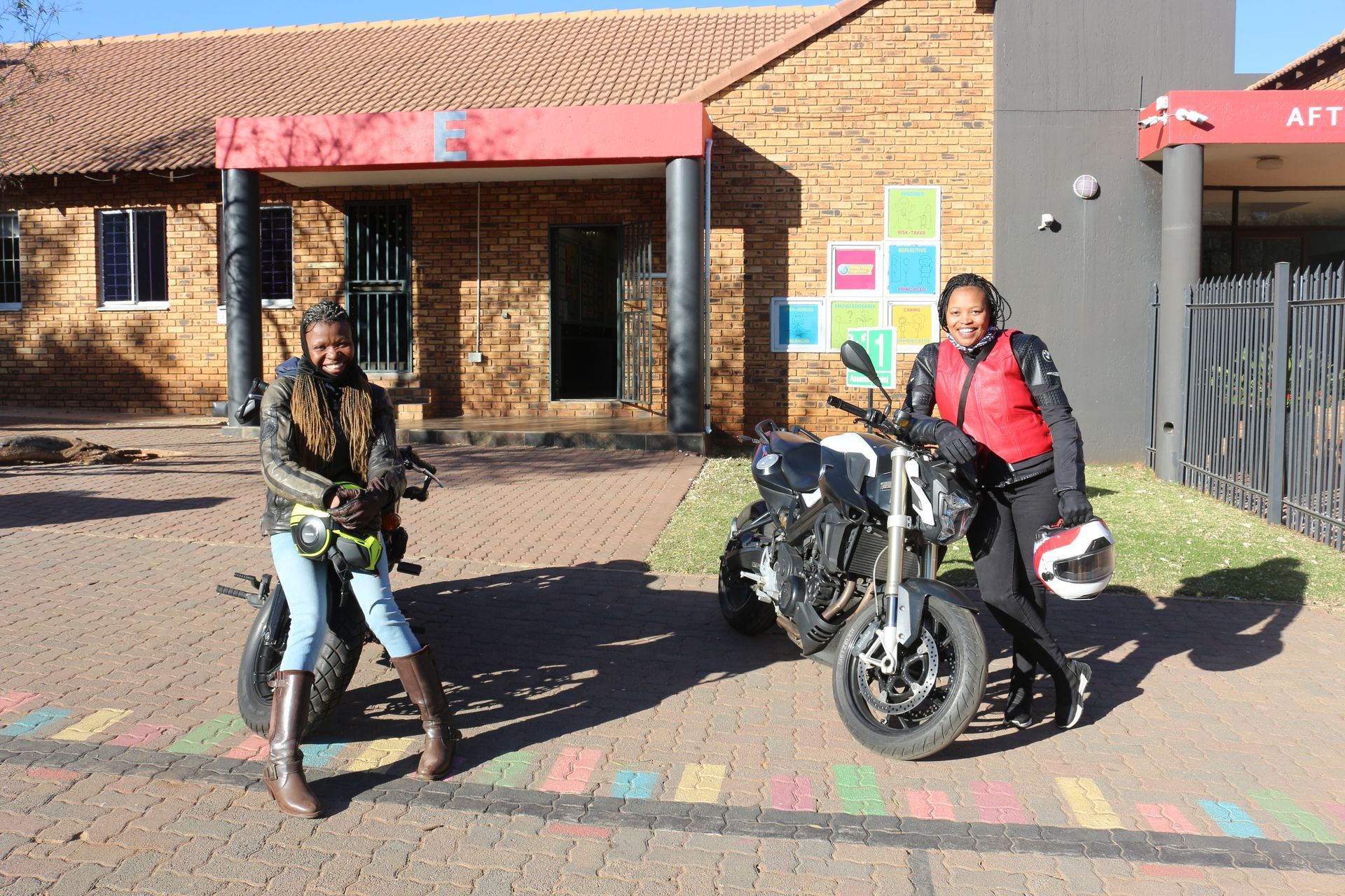 Two women are standing next to their motorcycles in front of a building.