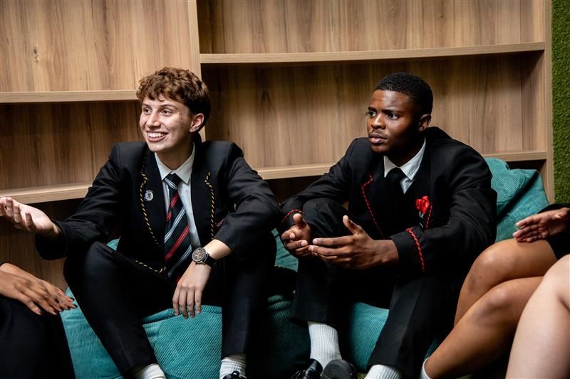 Two people in school uniforms sit on a teal couch, talking. Wooden wall backdrop.
