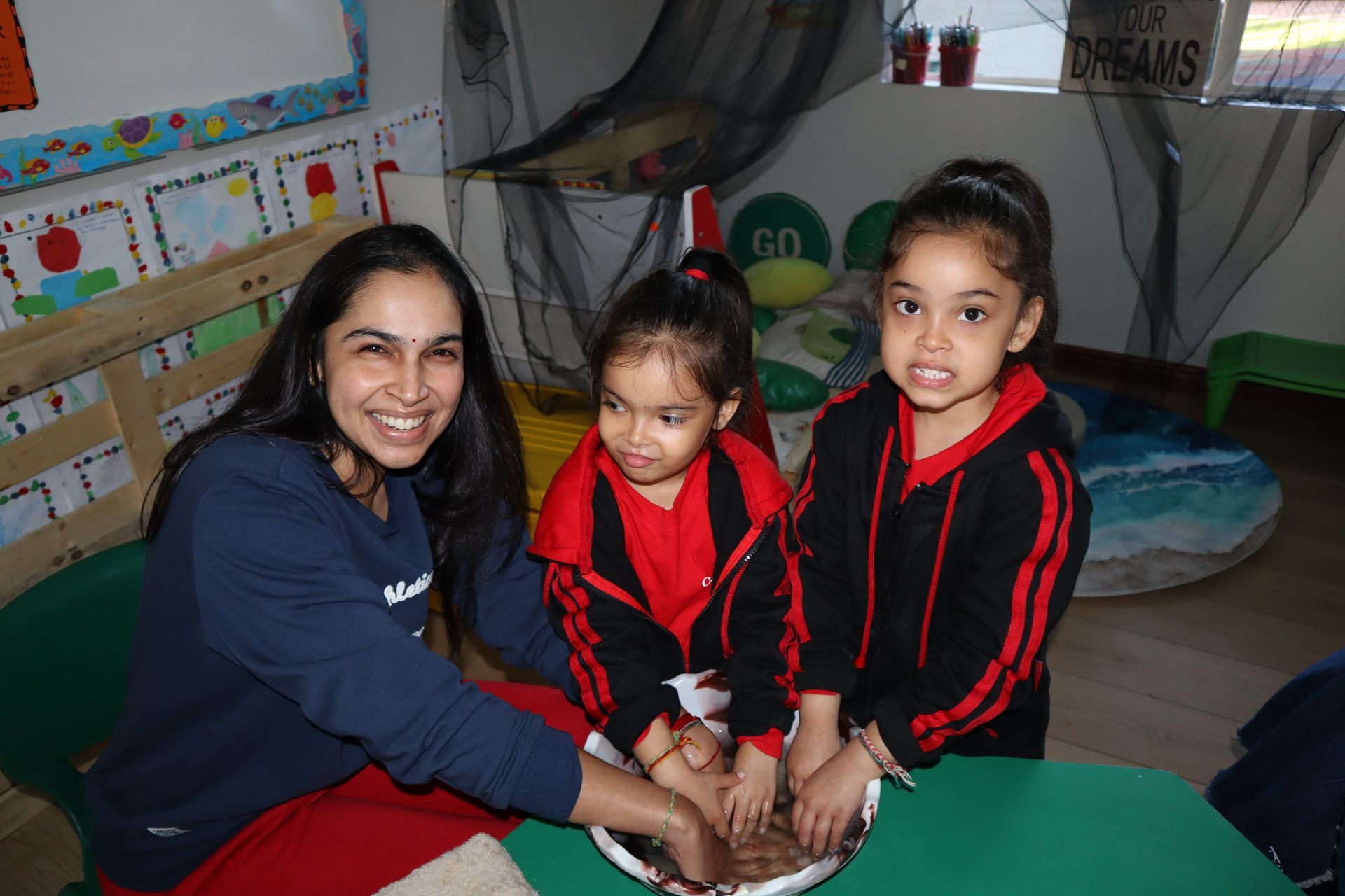 Woman and two children with hands in a bowl, smiling. Indoors, bright colors.