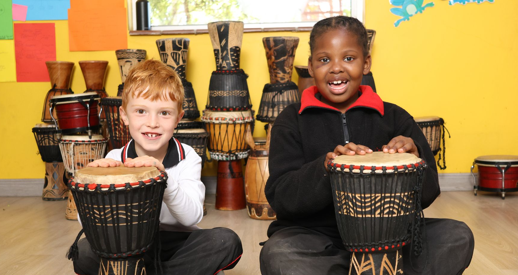 Two children smiling, sitting with drums. A room filled with drums, colorful walls.