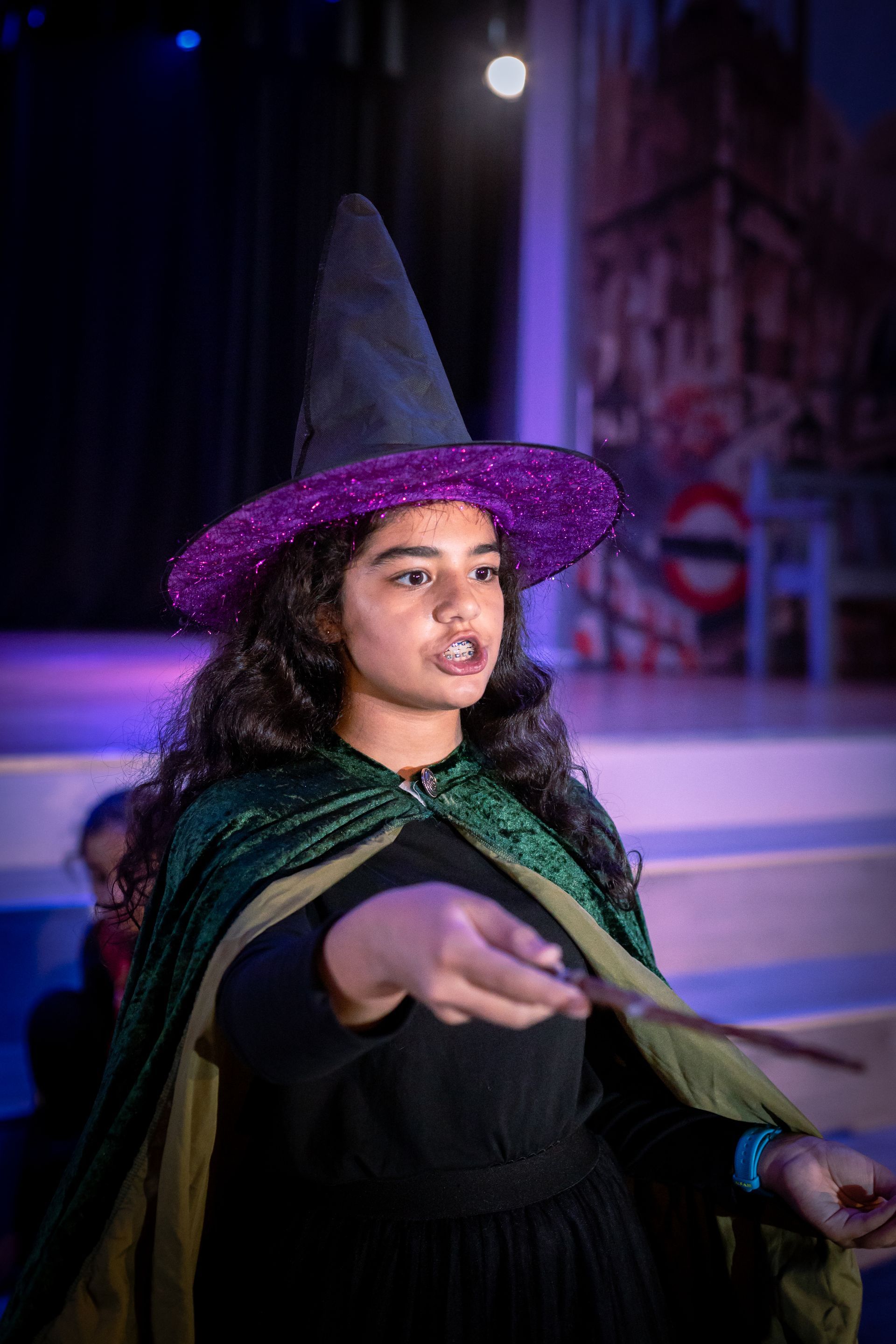 A young girl dressed as a witch is holding a wand on a stage.