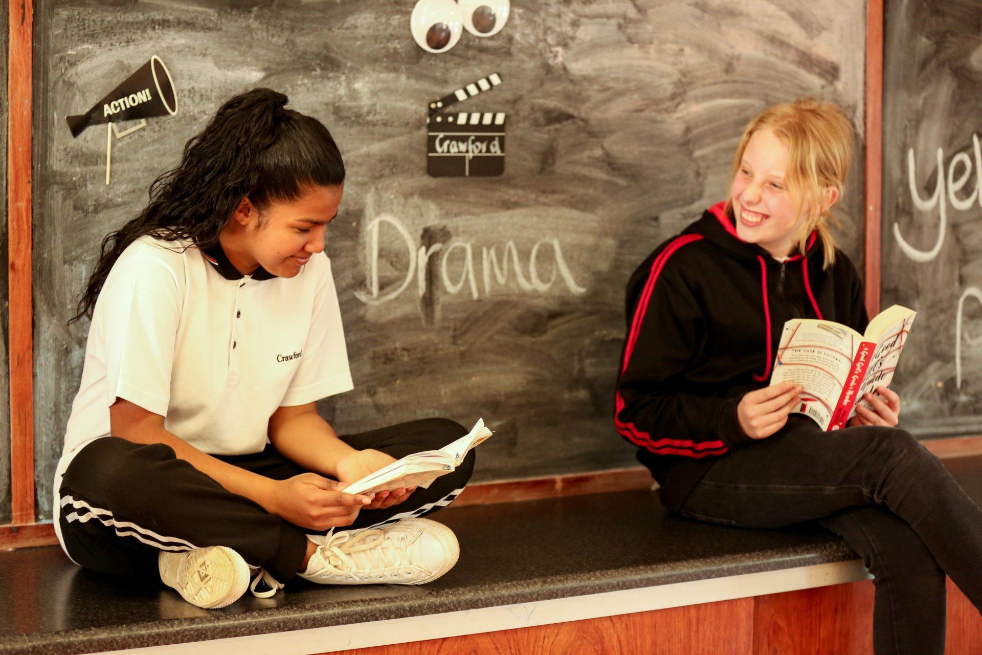two girls are sitting on a bench reading books in front of a blackboard that says drama