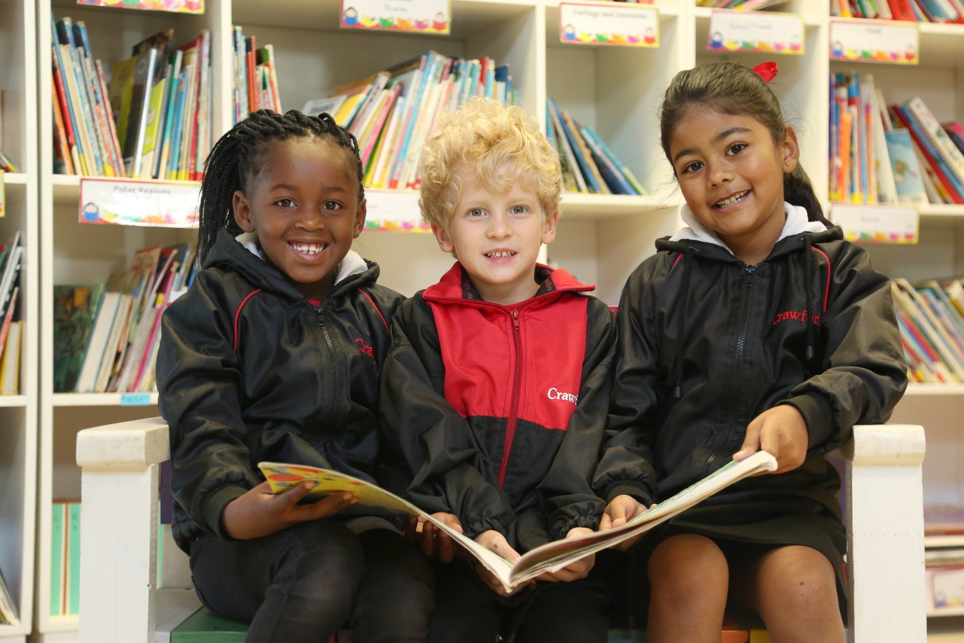 Three children are sitting on a bench reading books in a library.