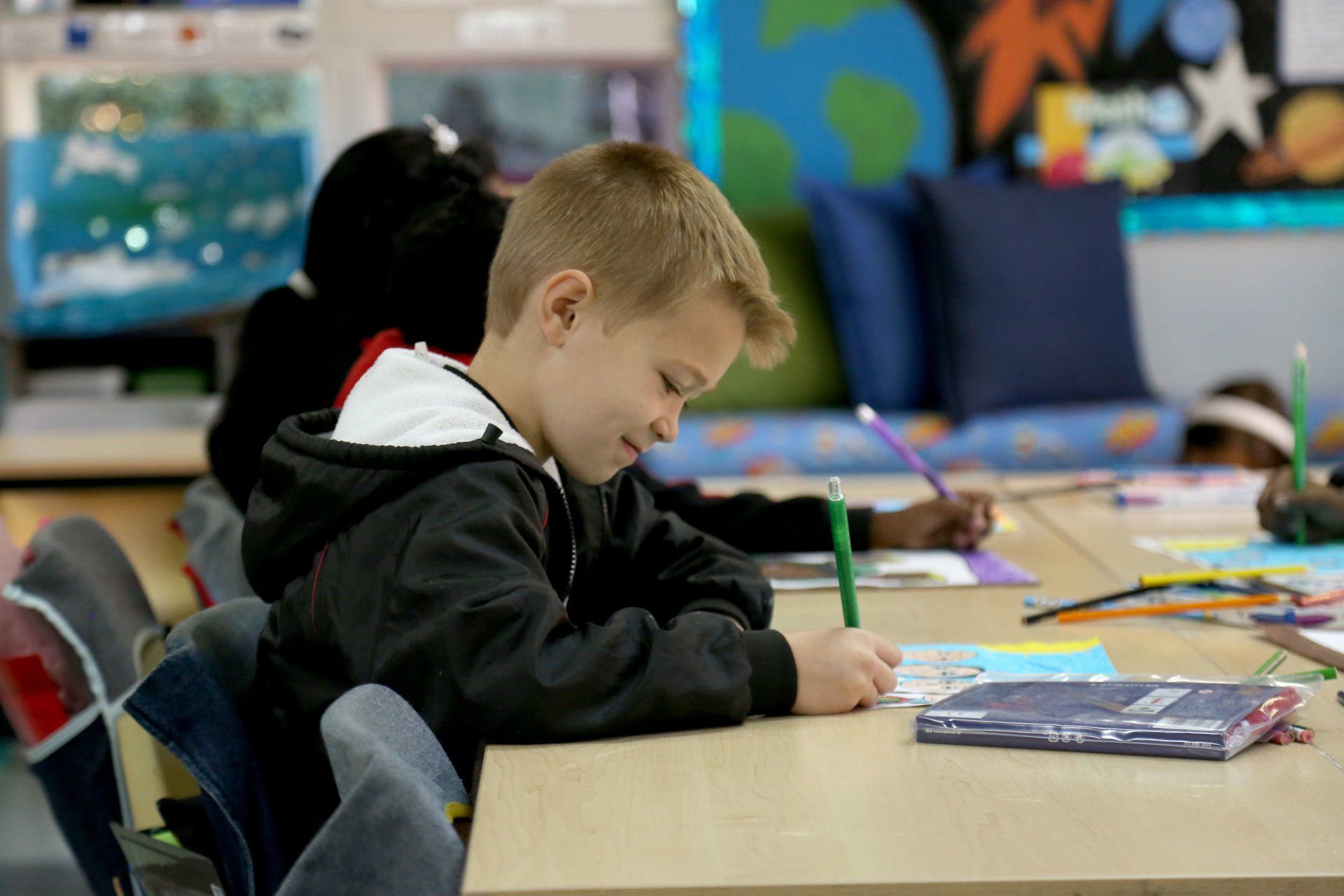 a young boy is sitting at a desk in a classroom writing on a piece of paper .