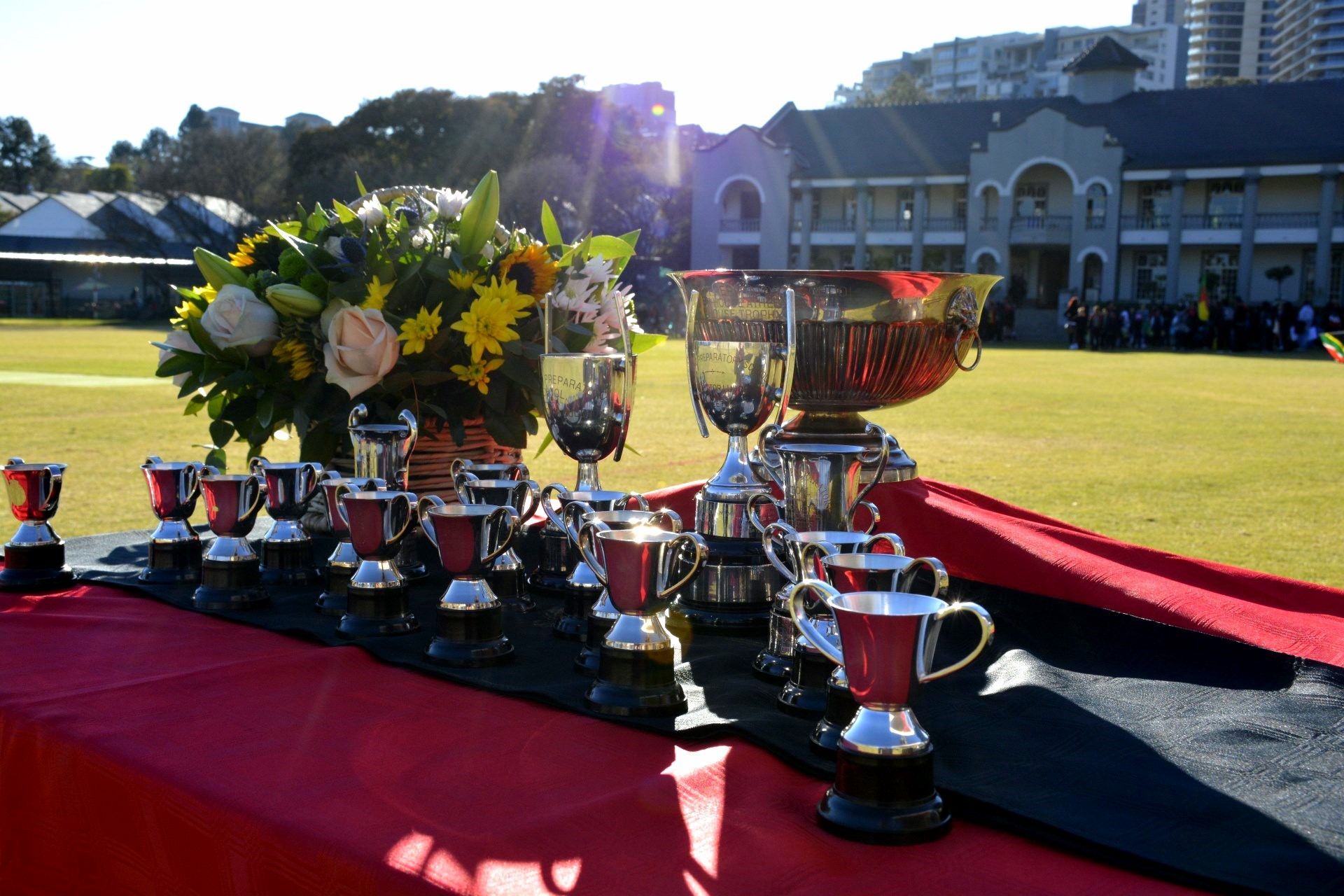 Trophies on a table with a red and black tablecloth, in front of a building and field with flowers.
