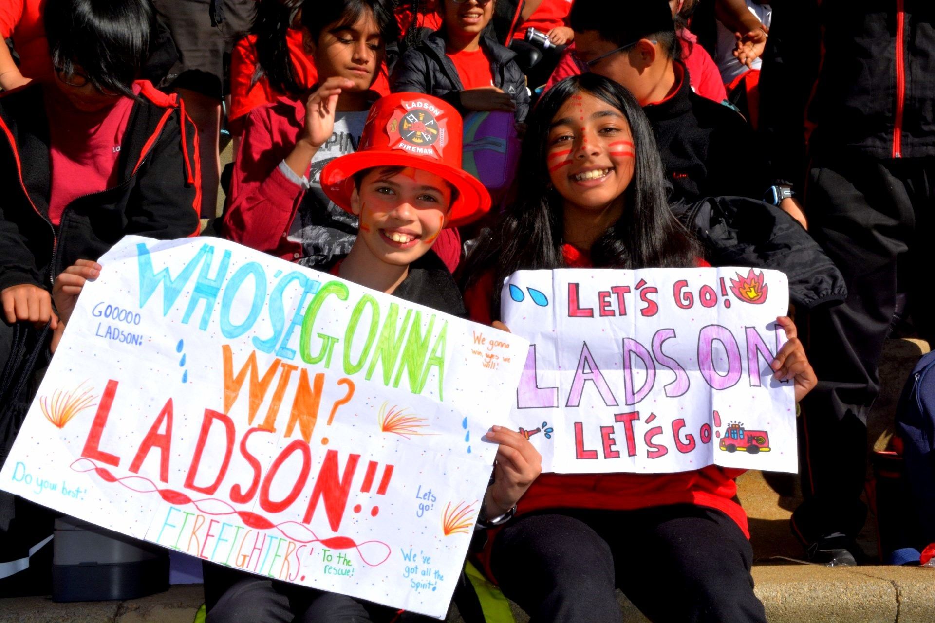 Two students hold signs at an event, one wearing a firefighter hat. Signs read