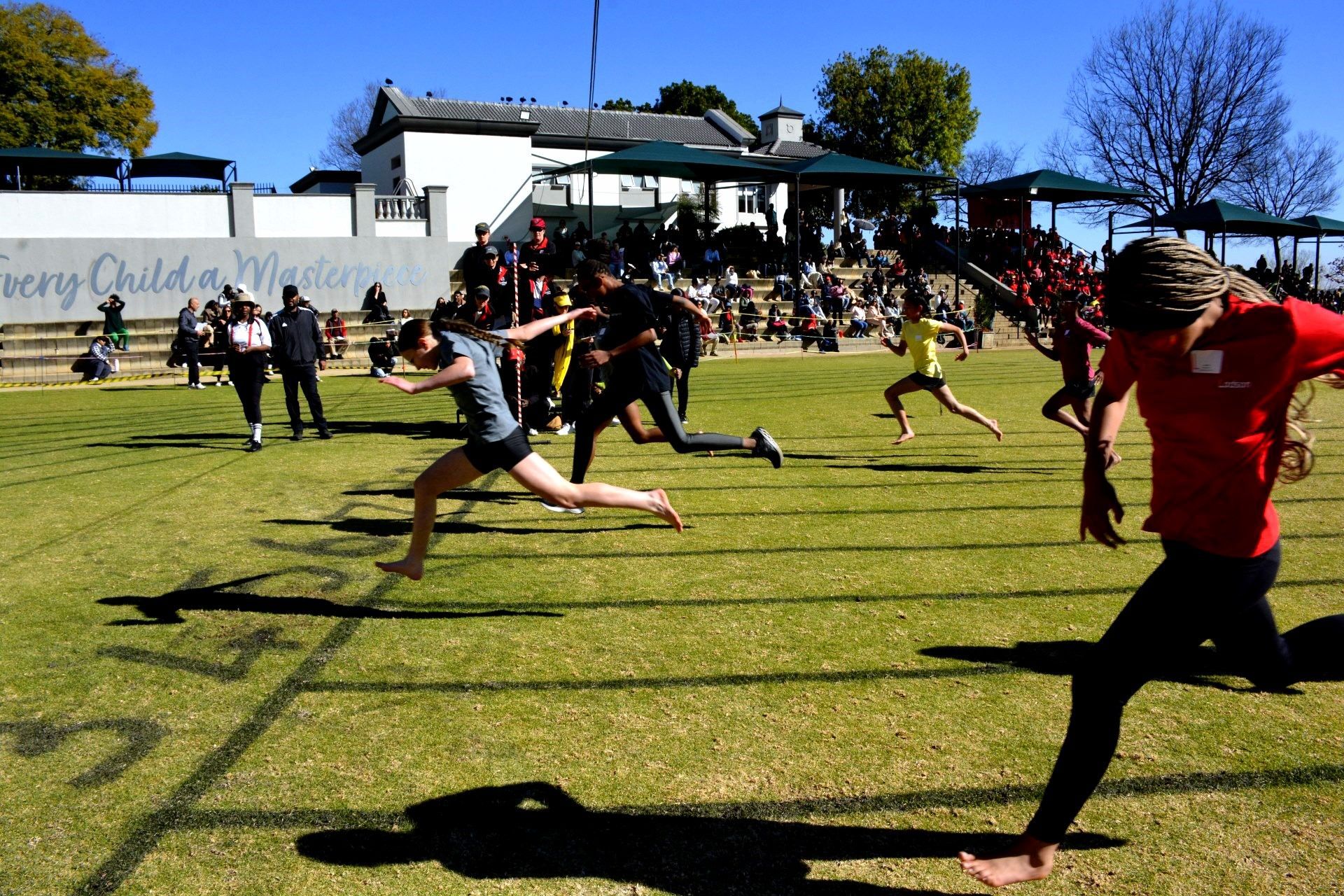 People running a track race on a grass field in front of a building with spectators.