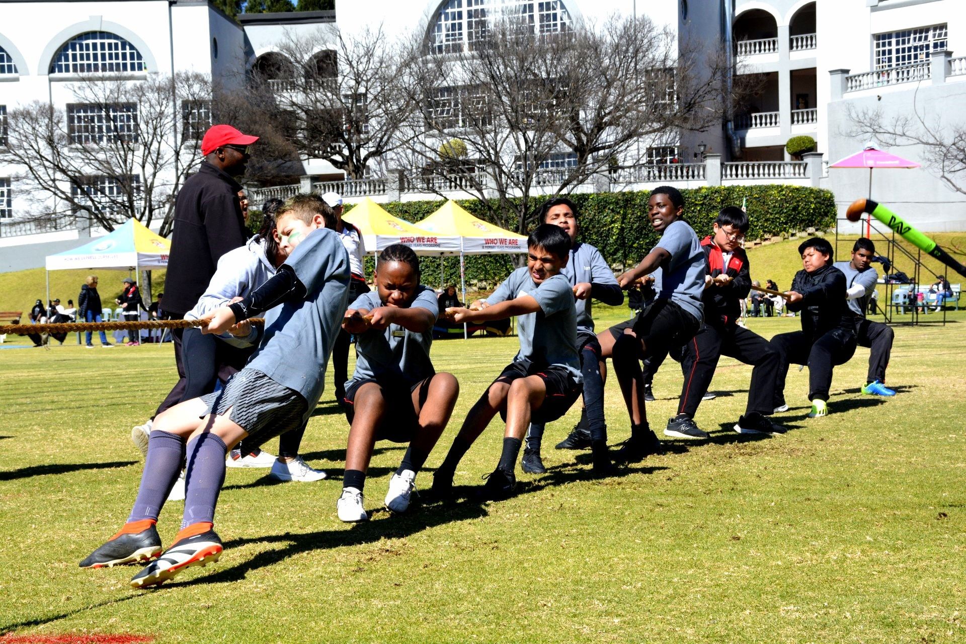 Children playing tug-of-war on a green field. A man in red hat coaches. White building in the background.