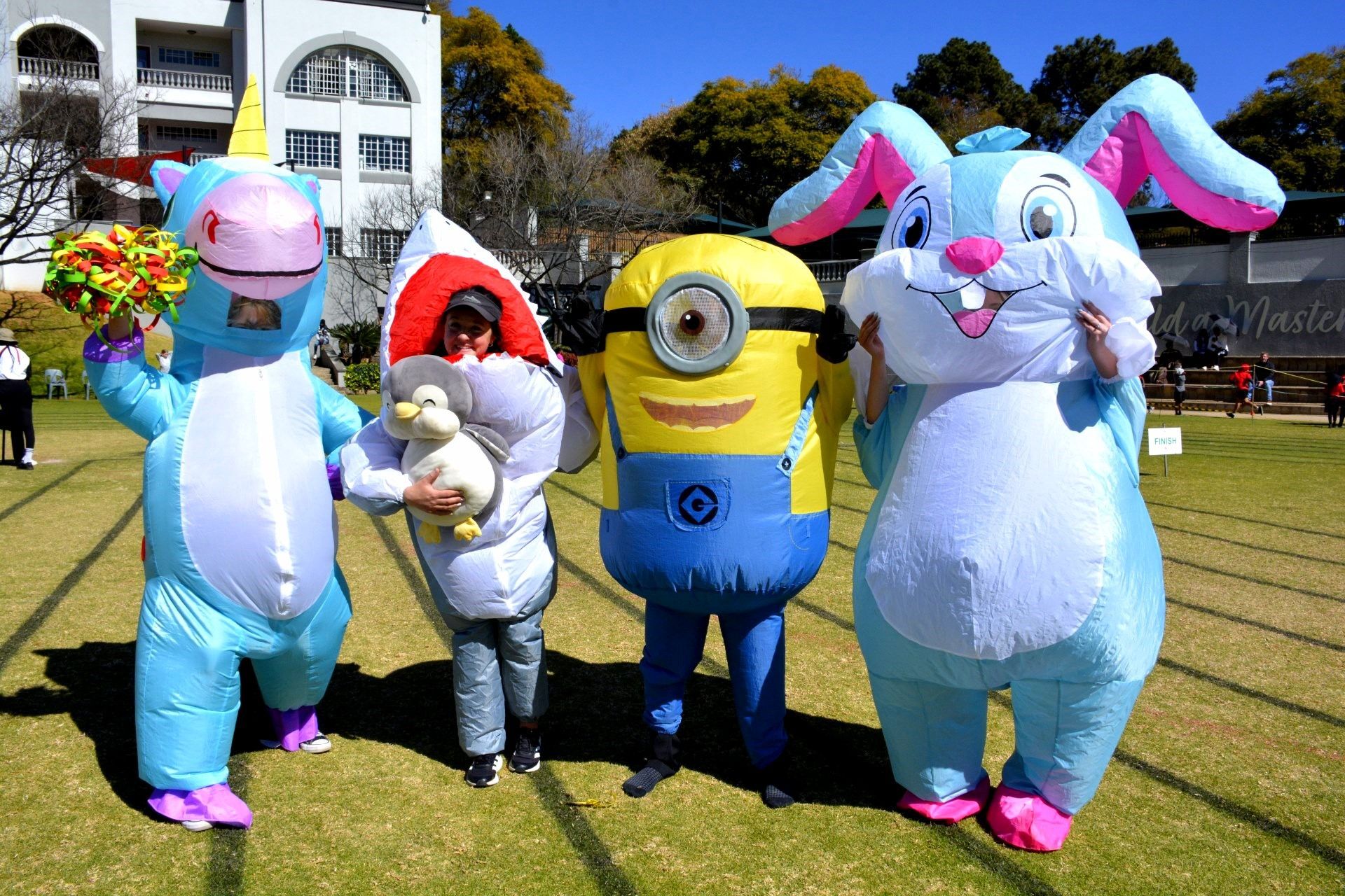 Four people in inflatable costumes: unicorn, shark, minion, and Easter bunny, on a sunny field.