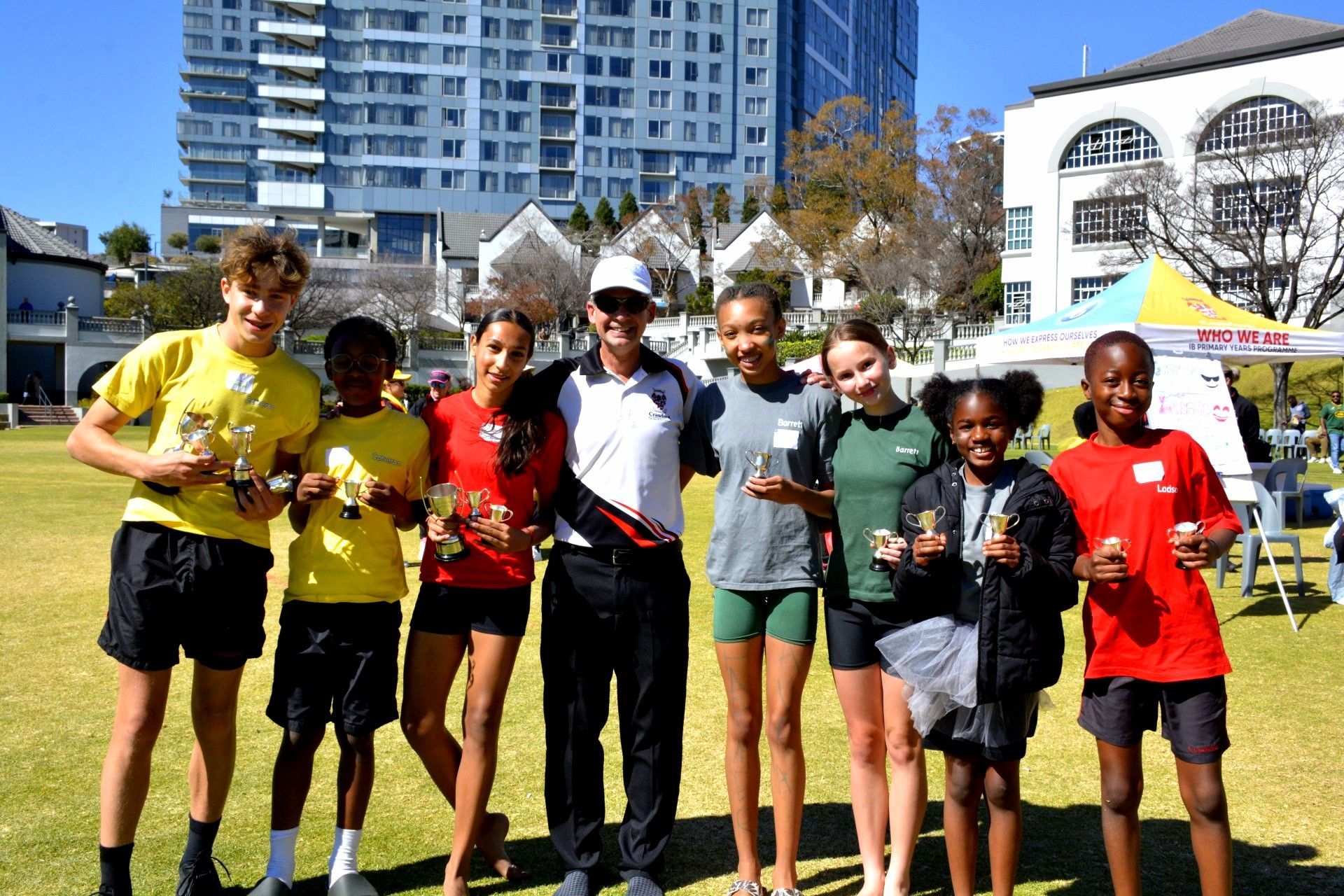 Group of runners holding trophies with a coach on a grassy field in front of a building.